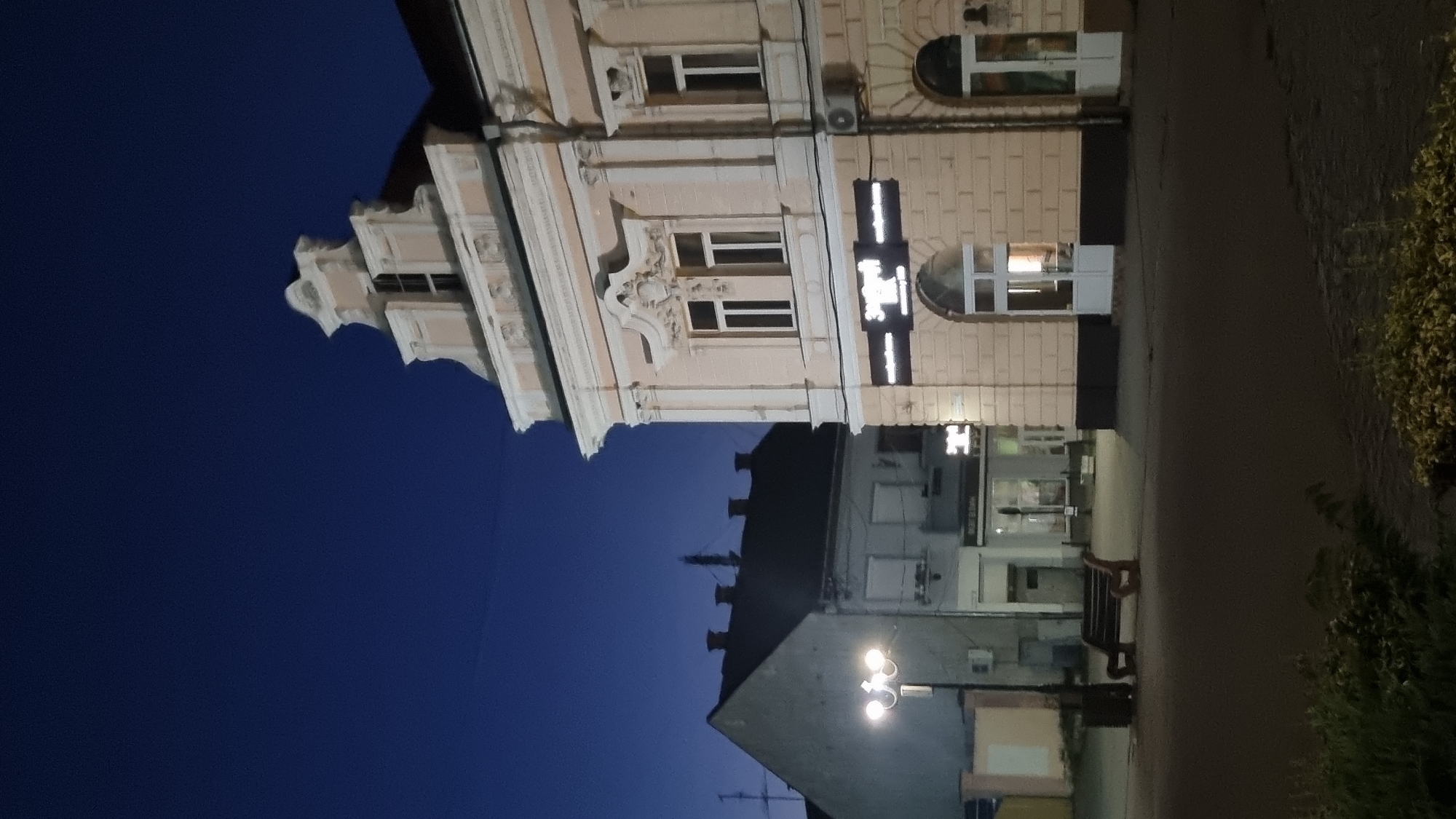 The image shows a street scene in Berehove, Ukraine, at twilight or night. A light-colored building with ornate architectural details, including decorative molding and a stepped gable, is prominently featured on the right side of the frame. This building appears to be a commercial establishment, with a lit sign above arched doorways. To the left of this building, a darker, sloped-roof building with multiple chimneys is visible, suggesting it is an older residential or utility structure. In the foreground, a street lamp illuminates a park bench, and a dark, paved road or plaza stretches across the lower part of the image. The sky is a deep, clear blue, indicative of dusk or early night. The overall impression is of a quiet urban street at the end of the day.