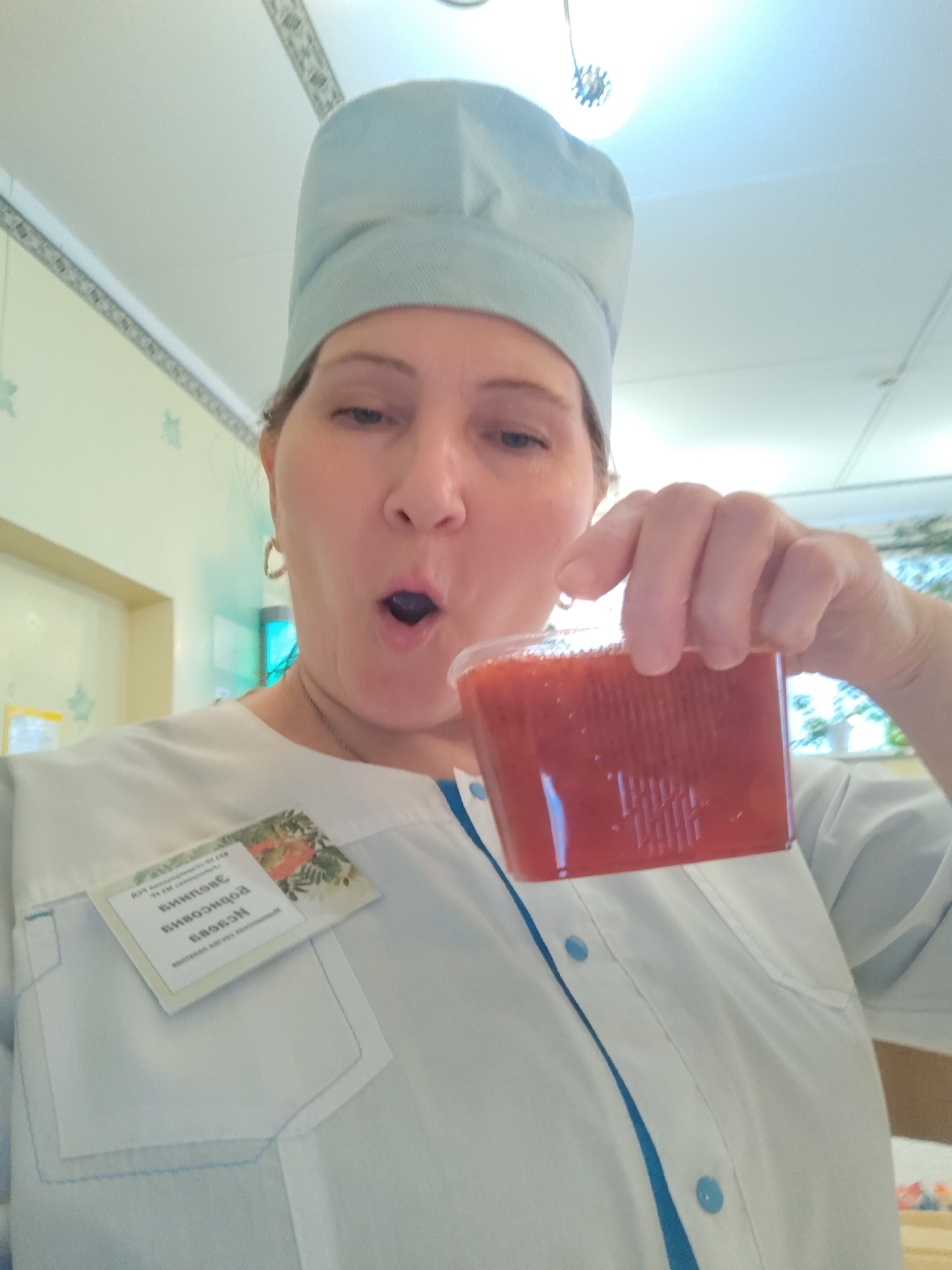 A woman, seemingly taking a selfie, is pictured from the chest up, holding a clear plastic cup filled with a bright red or orange liquid. She wears a light blue scrub cap and a white uniform shirt featuring blue trim and buttons, indicative of a professional, likely medical or service, role. A gold hoop earring is visible on her left ear. Her mouth is open in an expressive, perhaps surprised or anticipatory, gesture, as if about to drink from the cup.
Pinned to her uniform, near a pocket, is a reversed name tag with Cyrillic text. The visible text reads "ВЫДАНО ЗАМ" (Issued to Deputy/Assistant) followed by the name "РОДИОНОВОЙ НАТАЛЬЕЙ" (Rodionova Natalya). The tag also includes a small graphic of red berries or flowers on a green background.
The setting appears to be an indoor institutional environment, possibly a clinic, hospital, or cafeteria located in Sarapul, Russia. The background features a ceiling with a decorative border and a modern light fixture. Faint light green leafy or floral patterns adorn the wall. A blue-glowing object, possibly a decorative light or a sanitization device, is also visible. The general lighting suggests it is daytime. The woman's open-mouthed expression conveys a sense of playfulness or excitement.
