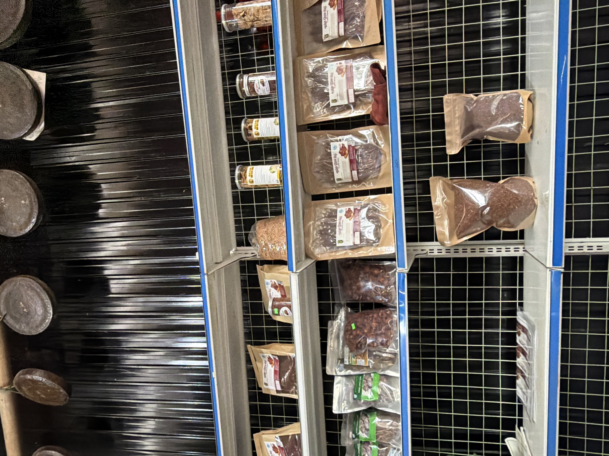 This image captures an indoor retail display, likely in a shop or market in Măng Đen, Vietnam. The scene features multiple vertical shelving units filled with various packaged food items, set against a dark, grid-patterned background.
On the far left, partially visible, are several round, dark, rustic-looking objects, possibly coasters, small plates, or artisanal pieces. To their right are several shelves holding clear glass jars with screw-on lids, containing dried goods such as nuts, seeds, or dried fruits. Below these jars, and continuing down the shelves, are numerous flat, rectangular pouches, typically with a clear plastic front and a brown paper back. These pouches contain what appears to be dried meat, jerky, or possibly preserved fruits, and each has a white label featuring illustrations and text, likely in Vietnamese script. One label prominently displays "Mứt..." which translates to preserved fruit or jam.
Further to the right, another set of shelves holds similar packaged goods. Many are clear plastic pouches containing reddish-brown, chunky items that resemble dried fruit, nuts, or possibly coffee beans. Below these, some pouches feature green and white labels. On the far right, two isolated, bulkier clear plastic pouches contain a finer, granular reddish-brown substance, potentially ground coffee or a spice mix.
No people are visible, but the arrangement suggests a retail environment where customers would browse and purchase goods. The lighting appears to be artificial indoor lighting. The visible packaging and the Vietnamese script on some labels confirm the location context of Măng Đen, Vietnam, indicating local produce or specialty items.