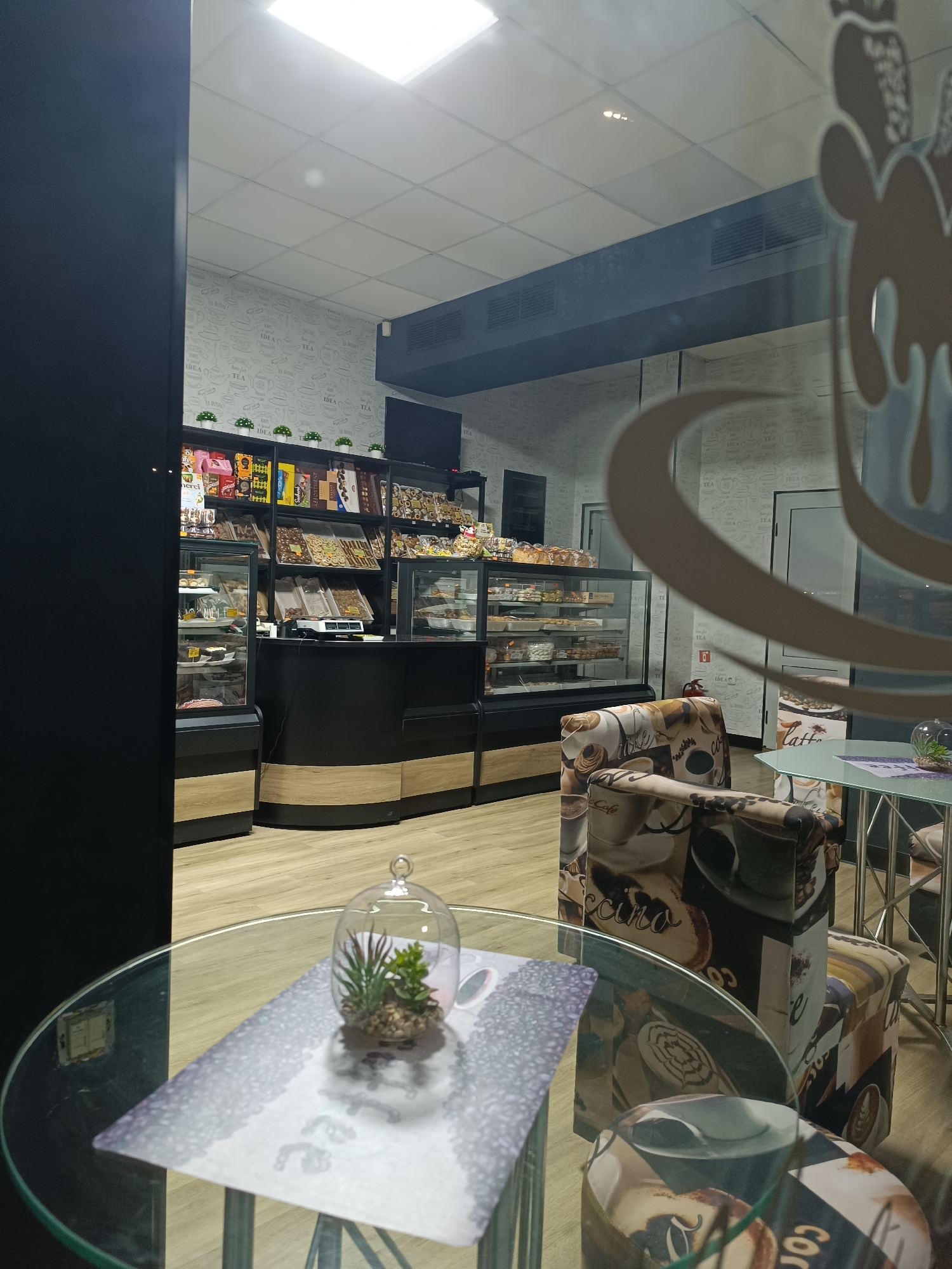 The image captures the interior of a cafe in Pleven, Bulgaria. Display cases filled with pastries and chocolates line the back wall, topped with shelves holding packaged goods and decorative plants. A black counter runs along the front of the display, creating a service area.
In the foreground, a glass-topped table holds a decorative terrarium with succulents. The table is set with a decorative placemat featuring coffee-themed designs. A chair upholstered in a similar coffee-themed pattern sits beside the table.
The overall aesthetic is modern and clean, with a light color palette and decorative accents. The cafe appears to be viewed through a window with a logo or design on it, partially obscuring the view.