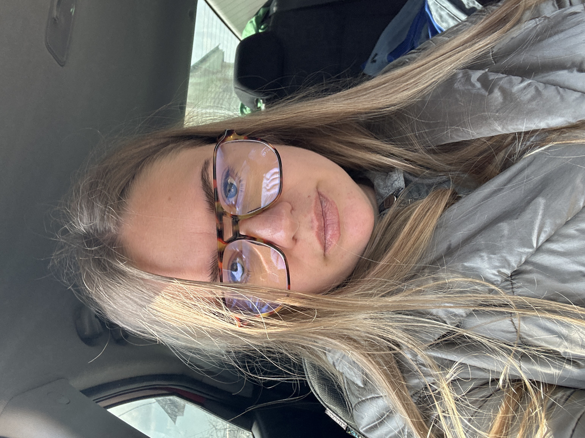 A woman with long blonde hair and glasses is sitting in a car. The car's interior is visible, including the dashboard and seats. The background shows an urban environment with buildings visible through the car window. The woman is wearing a grey jacket. The location is Уральское, Russia.
