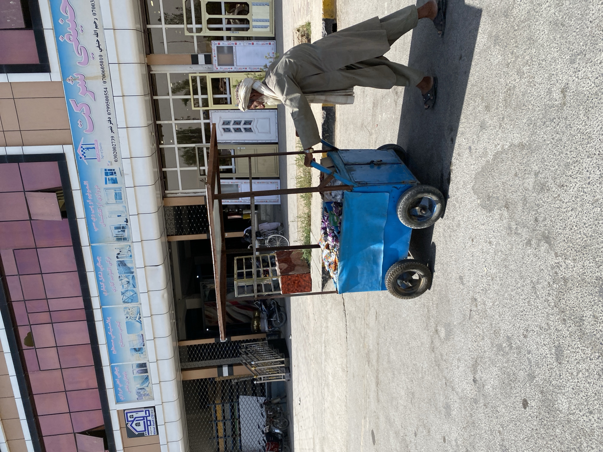 A vendor pushes a blue cart filled with various food items. The cart has two wheels at the front and two at the rear, and a canopy supported by metal poles. The vendor, an elderly man wearing a traditional shalwar kameez and a turban, is pushing the cart with his right hand while his left arm is extended. He is walking on a paved street in front of a building. The building has a modern facade with tinted windows and a sign in Arabic script that reads "Hameefi Company". The sign also contains smaller text with contact information, including phone numbers. The ground is a rough, light-colored asphalt. In the background, several motorcycles and a bicycle are parked. The scene appears to be a street in Kandahar, Afghanistan, given the architectural style and signage.