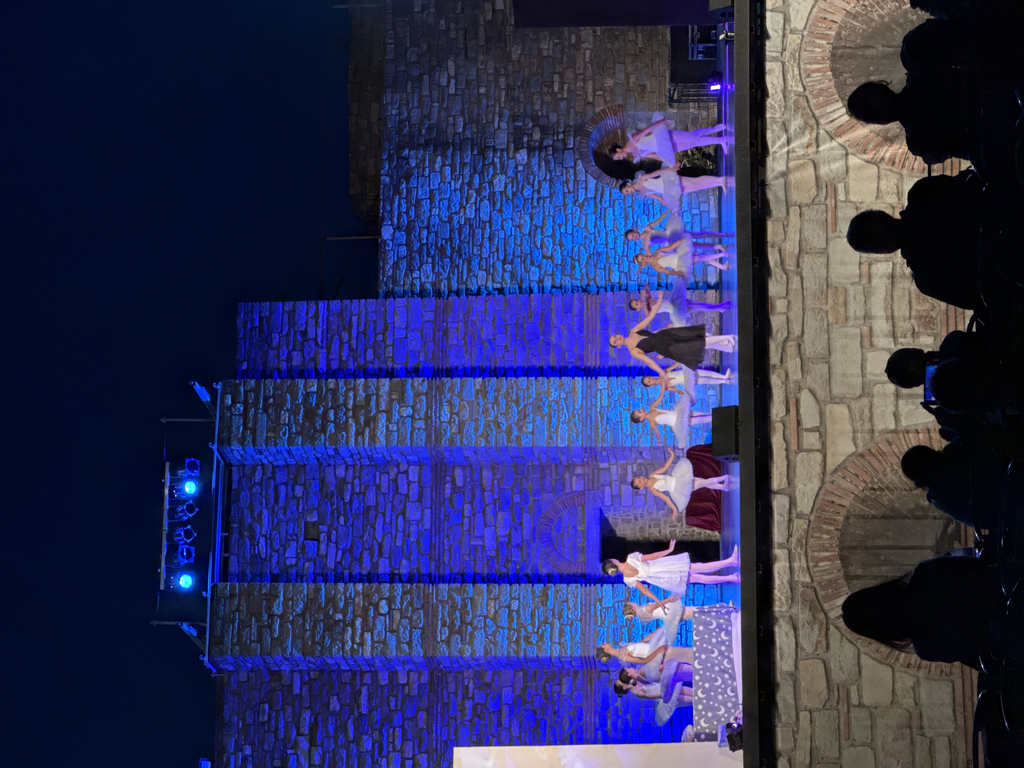 A group of young ballerinas in white tutus perform on an outdoor stage at night, bathed in bright blue light. A woman in a black dress, likely their instructor, stands to the left of the group, observing them. The stage is set against a backdrop of a stone wall, illuminated by more blue lights, creating a dramatic effect. In the foreground, the silhouettes of audience members are visible, seated in dark chairs, facing the performance. The scene appears to be taking place in Veliko Tarnovo, Bulgaria, possibly within the Tsarevets Fortress, known for its historical significance and nighttime performances.