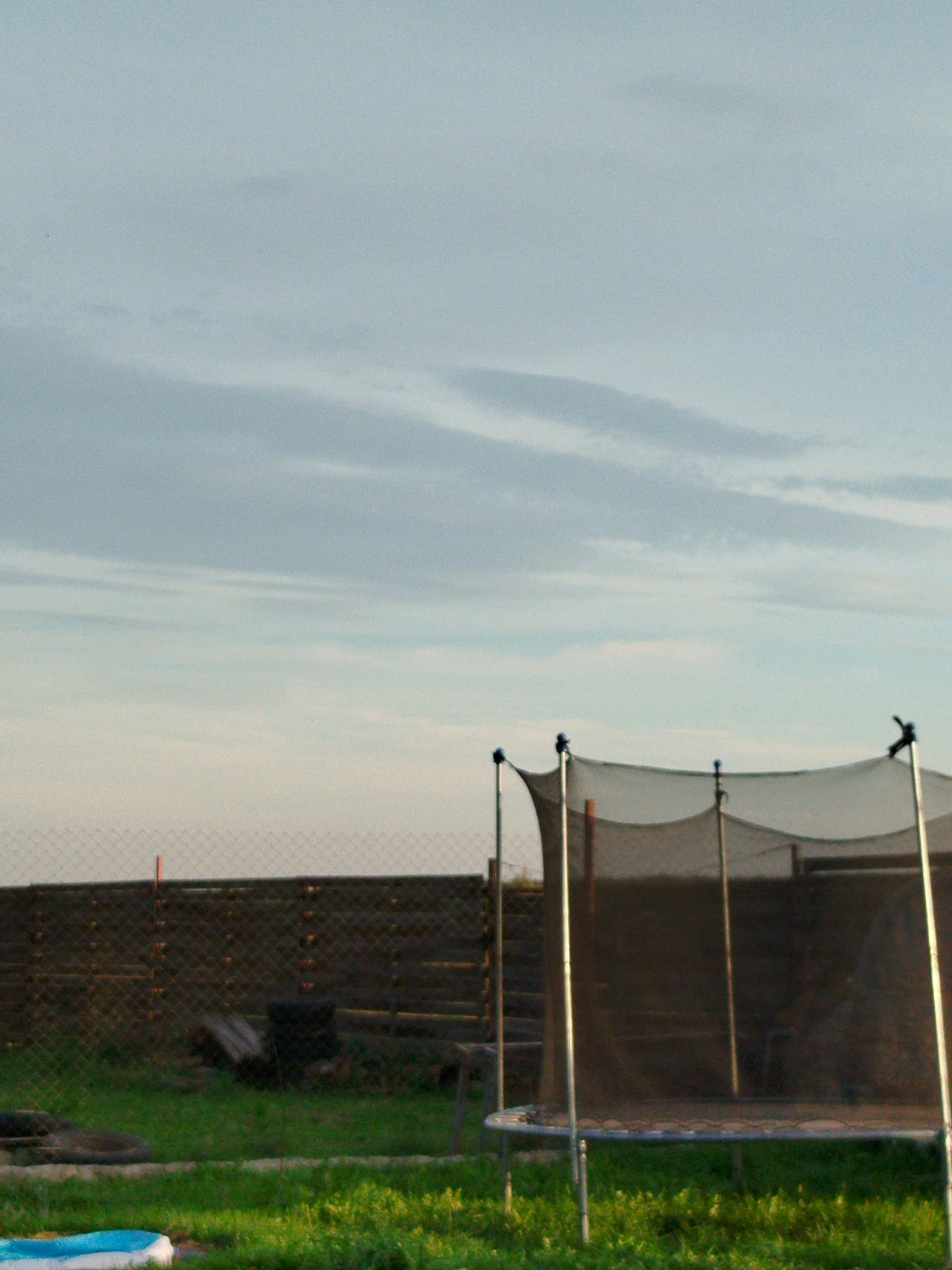 The image depicts an outdoor scene with a trampoline in the foreground, partially obscuring a wooden fence and a chain-link fence behind it. The trampoline has a black safety net and a silver frame. Beyond the fences, a grassy area is visible with some scattered objects, including what appears to be a black tire and some wooden planks. A small, blue and white inflatable pool is partially visible in the bottom left corner of the image. The sky is overcast with thin, wispy clouds. The overall impression is of a backyard or a rural property. No people are present in the image. The location is identified as Крывасельскі сельскі Савет, Belarus.