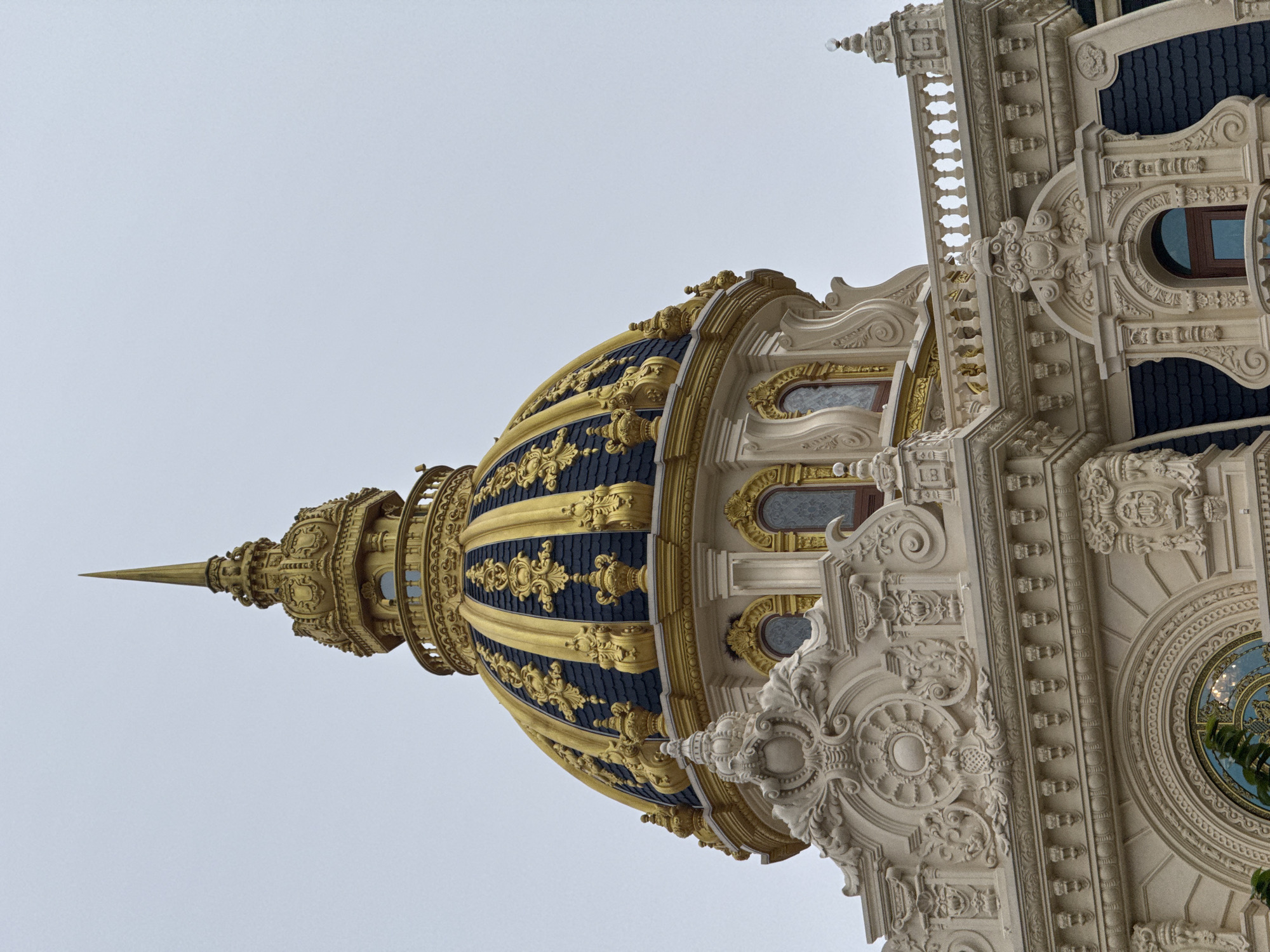 The image captures a close-up view of a gilded dome and ornate architectural details of a building in Da Lat, Vietnam. The dome is a prominent feature, adorned with intricate gold scrollwork against a dark blue background. It is topped by a tall, slender spire. Below the dome, the building exhibits elaborate cream-colored stucco ornamentation, including decorative moldings, reliefs, and arched windows. A balustrade runs along the edge of the visible rooftop. The sky is overcast and a pale gray, suggesting a cloudy day. The architectural style appears to be grand and possibly of European influence, a characteristic found in some of Da Lat's colonial-era structures. The building is the Dinh 1 Da Lat, also known as the Palace of Trai Mat.