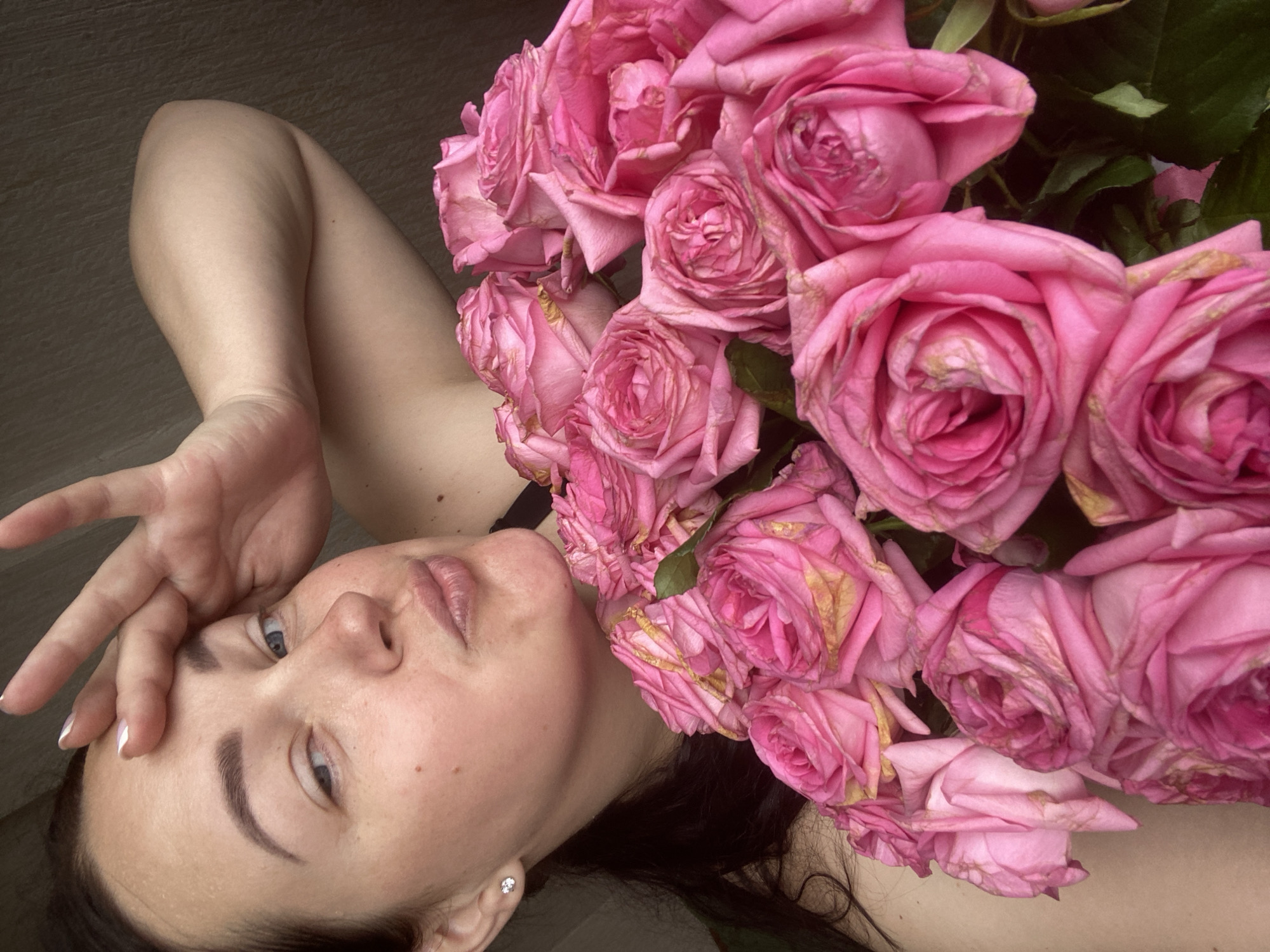A woman with dark hair and fair skin is lying down, holding a bouquet of pink roses. Her left hand is raised near her forehead. She is wearing a small earring in her left ear. The background is a plain, neutral color. The location is unknown, and there is no information to indicate it is in Sochi, Russia.
