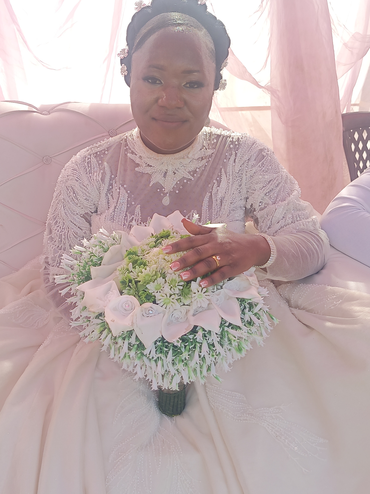 Akwanga, Nigeria: A woman sits on a white tufted couch, holding a bridal bouquet. The woman has dark hair styled up with floral accents. She wears a white, long-sleeved, lace-embellished gown with a high neckline. Her nails are manicured with light pink polish and white tips. She wears a gold ring and a diamond bracelet. The bouquet features white roses and small white and green filler flowers. Pink fabric drapes are in the background.