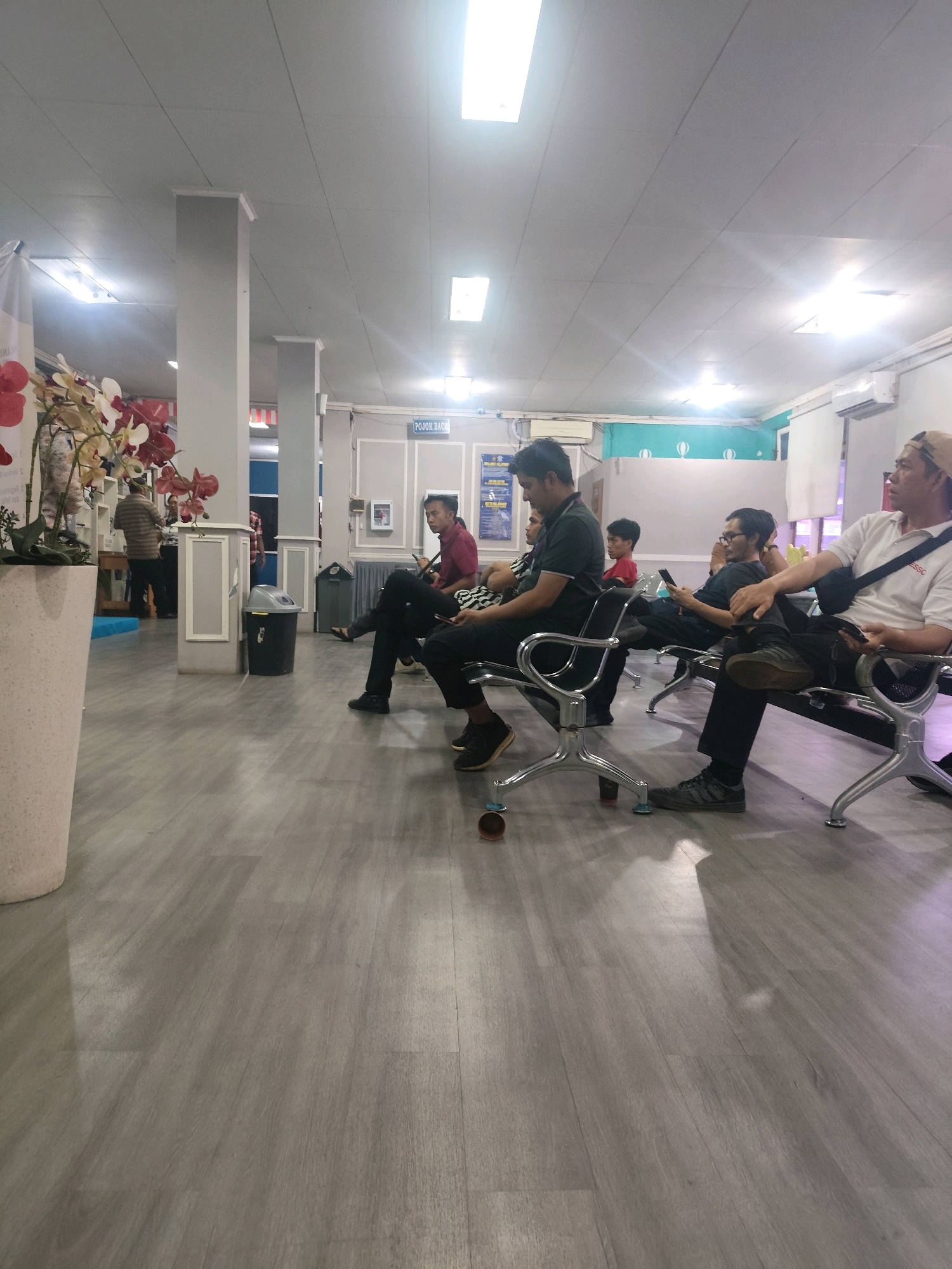 The image shows a waiting area with several people seated on chairs and a bench. The flooring is a light grey wood-look laminate. In the background, there are pillars, a trash can, a potted plant with artificial flowers, and signage indicating "POJOK BACA" (reading corner) and an air conditioner unit. The lighting appears to be fluorescent. It is plausible this scene is located within a public building in Palembang, Indonesia, such as a government office, clinic, or transportation terminal, given the signage and general waiting room atmosphere.