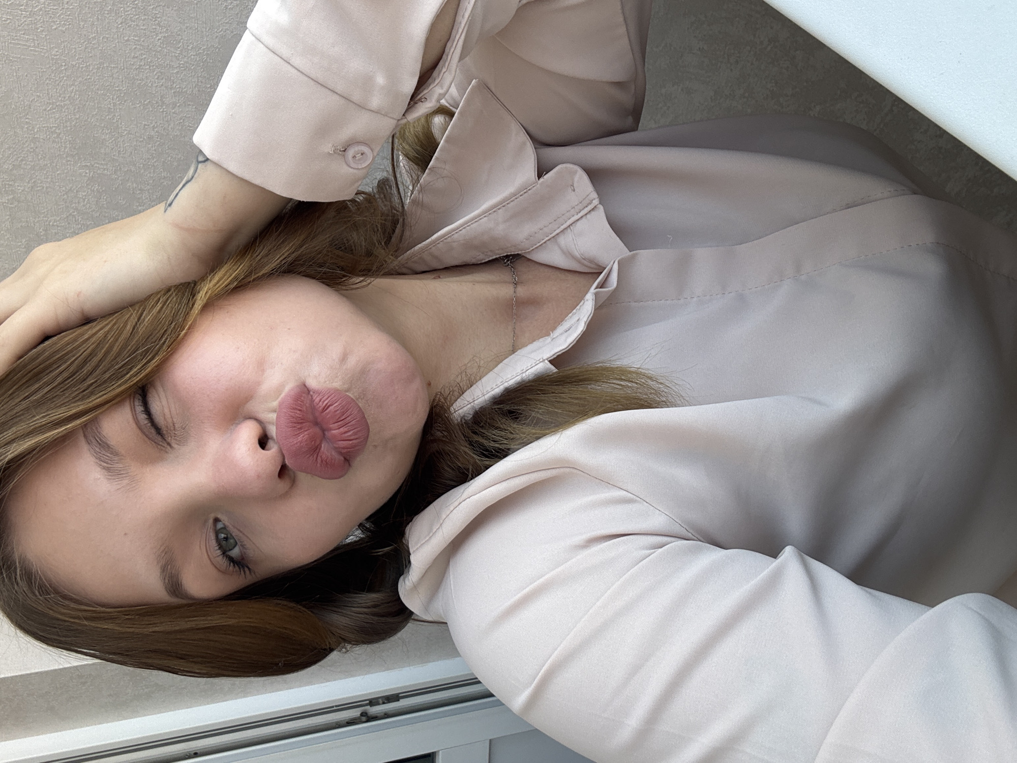 A young woman with light brown hair, wearing a light pink button-up shirt, is posing for a selfie. She is winking with her right eye and puckering her lips. Her left hand is touching her hair and head. A subtle tattoo is visible on her wrist. The background is a textured gray wall. She is wearing a delicate silver necklace.