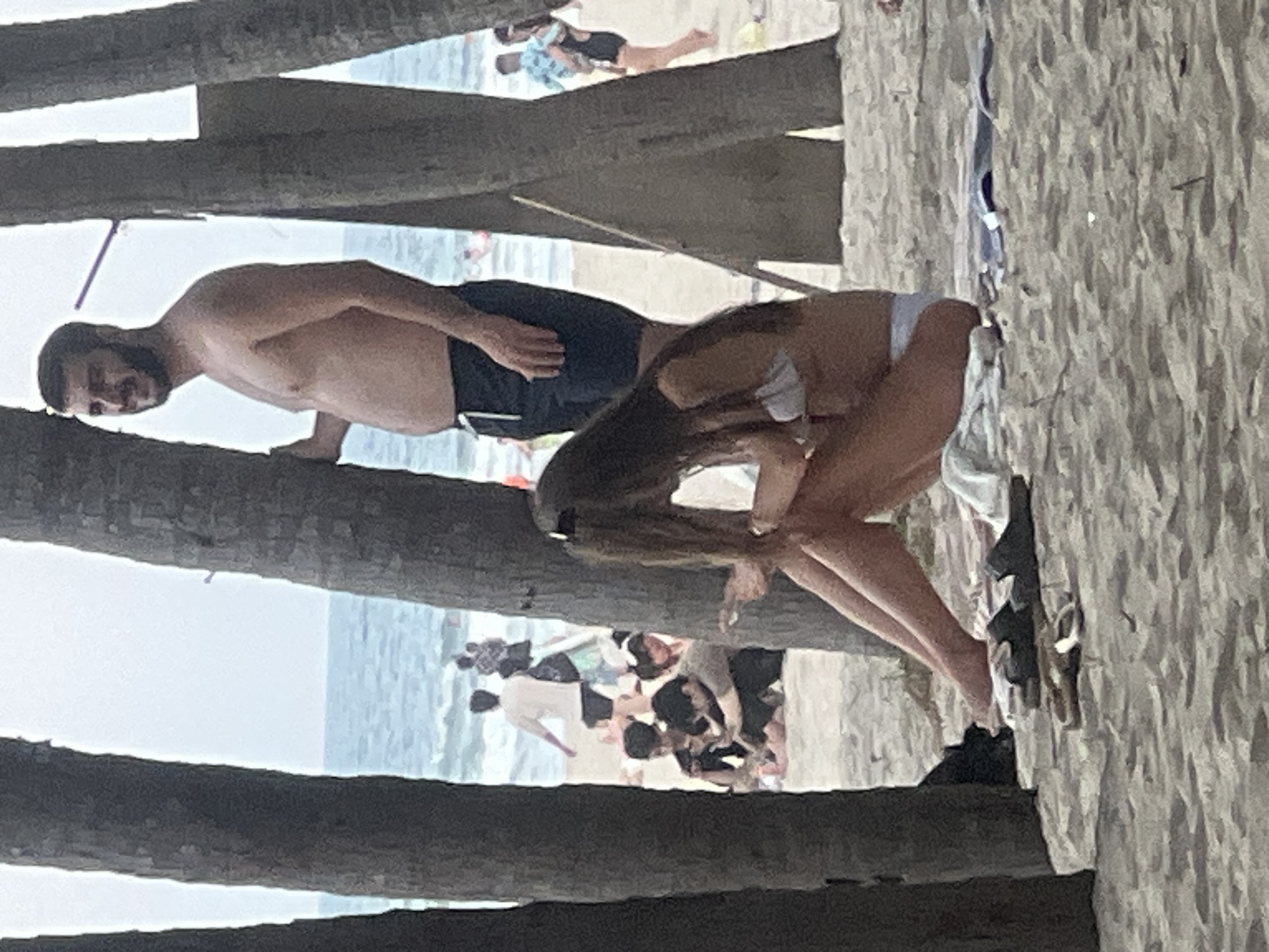 The image depicts a beach scene in Da Nang, Vietnam. In the foreground, a woman with long brown hair is sitting on the sand, wearing a white bikini top and bottom. She is looking down at her phone. Two pairs of sandals are on the sand in front of her. Behind her, a man with dark hair and a beard is standing, shirtless and wearing dark shorts. He is looking towards the sea. In the background, other beachgoers are visible, some walking and others sitting. The ocean is in the distance, with gentle waves. Palm trees frame the scene, with their trunks prominently featured in the foreground and midground. The sand is light brown and appears textured. The sky is overcast.