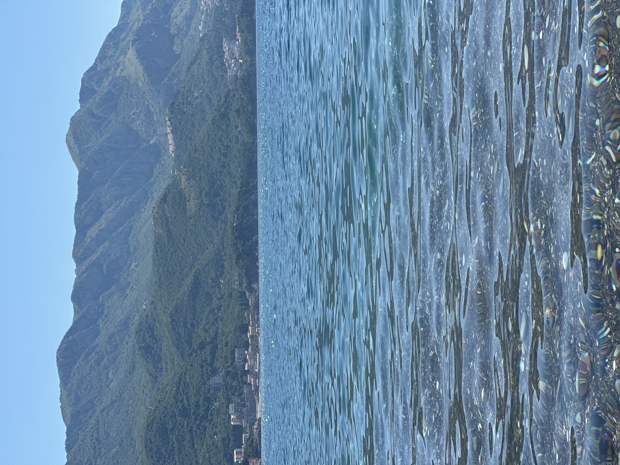 A horizontal image shows a portion of the Adriatic Sea and the adjacent landscape near Boreti, Montenegro. On the left, steep, forested mountains rise against a clear blue sky. At the base of the mountains, a small coastal settlement extends along the water's edge. The right side of the image is dominated by the turquoise water of the sea, with light reflecting off the surface and creating visible patterns of movement and ripple. No people or visible interactions are depicted.