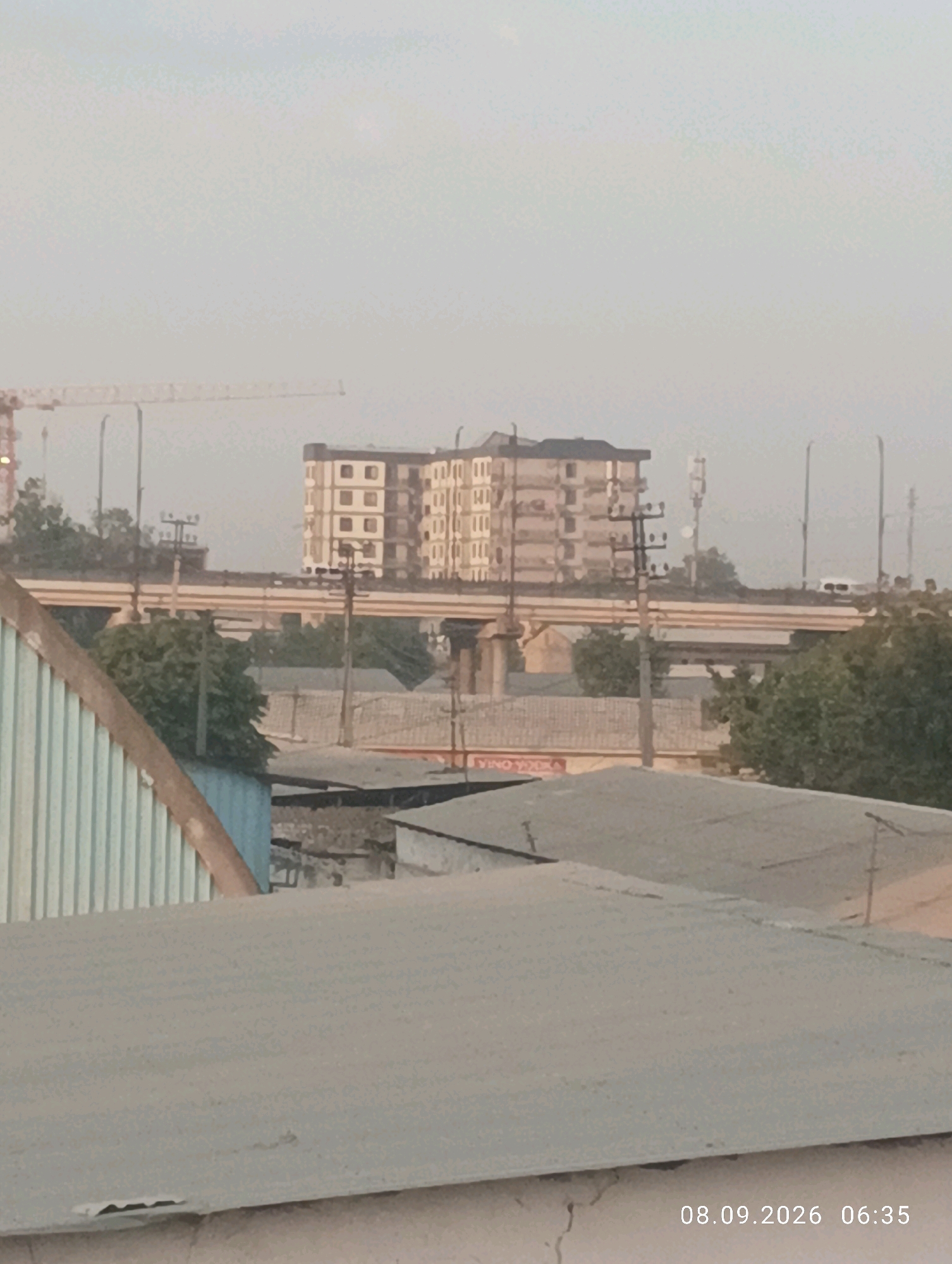 The image depicts a cityscape with several buildings, an elevated roadway, and a construction crane. In the foreground, there are several low-rise structures with corrugated metal roofs. In the middle ground, a multi-story apartment building with a repeating pattern of windows is visible, situated behind an elevated roadway. A construction crane is visible in the far left background. The scene appears to be of Yangi'ul, Uzbekistan, as indicated by the timestamp "08.09.2026 06:35" which suggests a date within Uzbekistan's time zone and the overall urban landscape. The sky is overcast, and there is a slight haze, suggesting it could be early morning or dusk. No people are clearly identifiable in the image.