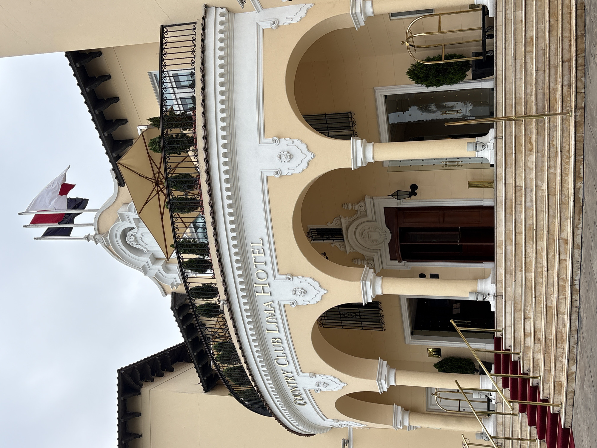 The image shows the entrance of the Country Club Lima Hotel, located in the San Isidro district of Lima, Peru. The building is a pale yellow, two-story structure with a curved facade featuring arches supported by columns. A wide, red carpeted staircase with golden railings leads up to the entrance. Above the arches, a white decorative band with the text "COUNTRY CLUB LIMA HOTEL" is visible. A black metal balcony with potted plants overlooks the entrance. The roof is tiled, and two flags are flying from a pole on the top of the building. The sky is overcast. There are no people visible in the image.