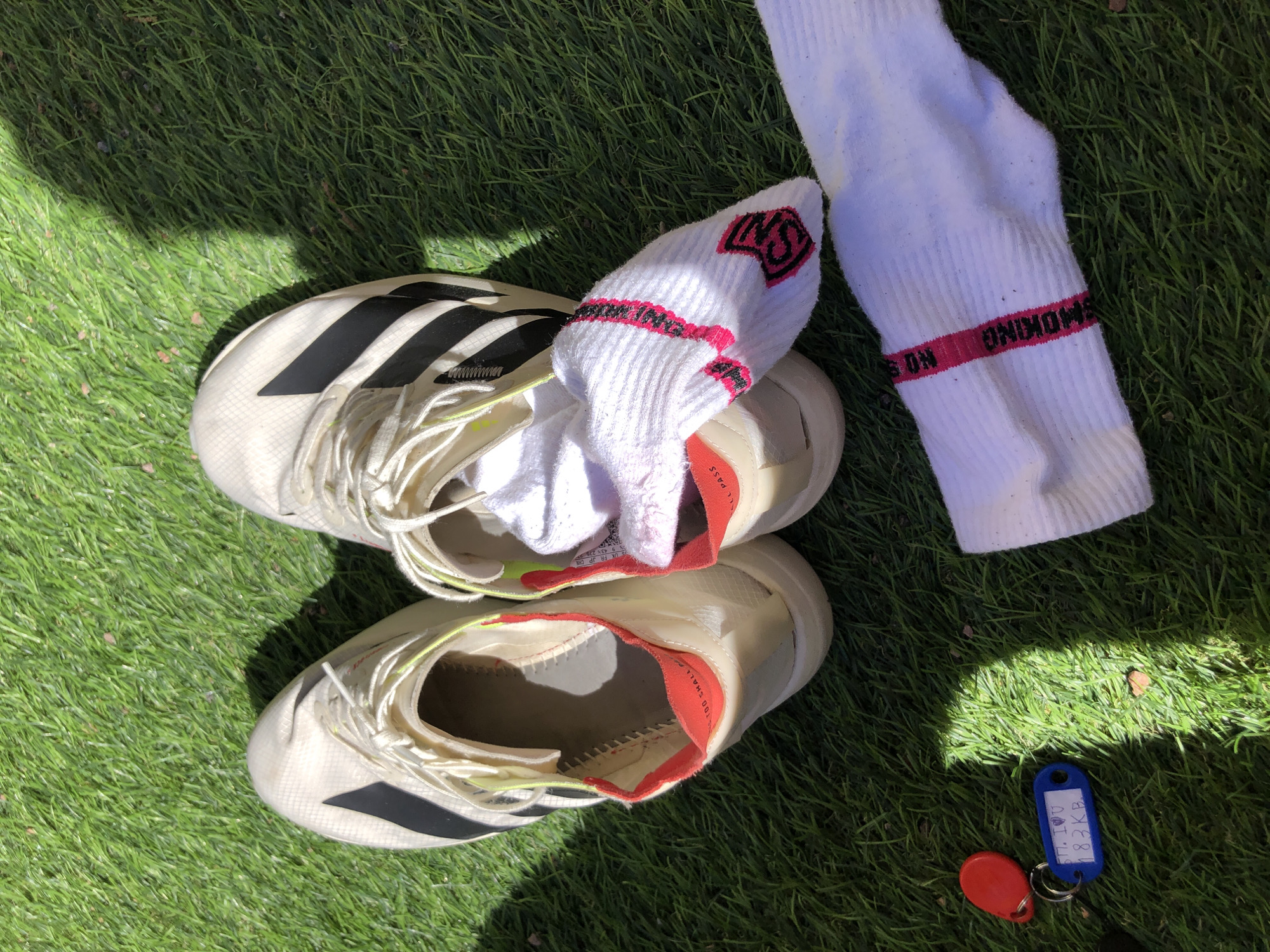 The image displays a pair of off-white Adidas athletic shoes with black stripes, two white socks with pink accents, and a set of keys, all resting on vibrant green artificial turf or well-maintained natural grass. The scene is brightly lit by direct sunlight, casting distinct shadows to the right of the objects.
The shoes appear to be worn, with untied laces and the right shoe containing one white sock. The left shoe is empty. Both shoes have a red/pinkish interior lining visible around the collar, with faint, partial text (possibly "TOO SHALL PASS") along the rim. A small Adidas tag is visible inside the right shoe.
One white sock is crumpled inside the right shoe, displaying a pink logo or emblem. The other white sock lies extended to the right of the shoes, showing a clear pink band with the text "SMOKING NO" printed on it, inverted relative to the top of the sock.
In the bottom left corner of the frame, a set of keys is visible. It includes a red, oval-shaped fob and a blue, rectangular fob attached to a metal ring. The blue fob has handwritten text: "БН. I ❤️U 183KB." (БН are Cyrillic letters, "I ❤️U" means "I love you," and "183KB" is an alphanumeric code).
Based on the key fob's text "БН", which is a common abbreviation for "Большая Не́вка" (Bolshaya Nevka), an arm of the Neva River in Saint Petersburg, Russia, and the athletic context (running shoes, sports socks, grass), the location is likely near a sports facility or recreational area along the Bolshaya Nevka, possibly on or around Krestovsky Island, which is known for its extensive sporting infrastructure in Saint Petersburg. The "183KB" on the fob may refer to a specific locker, unit, or address within such a complex.