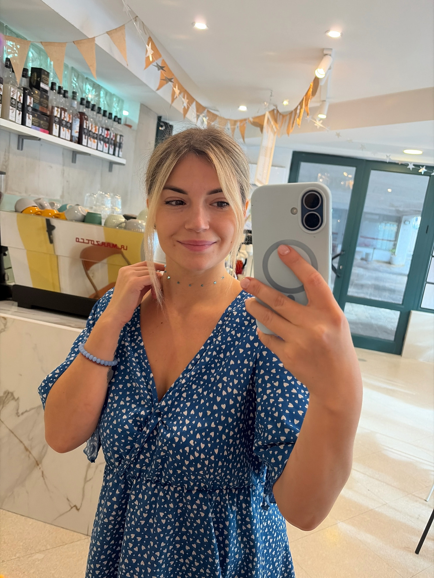 A woman with blonde hair pulled back and bangs framing her face takes a selfie in a brightly lit cafe in Budva, Montenegro. She is wearing a blue dress with a white heart pattern and a blue beaded bracelet on her left wrist, as well as a delicate choker necklace. Her right hand holds a light blue smartphone up to take the picture. Behind her, shelves are stocked with bottles of syrups and coffee cups are stacked on a counter. A yellow and orange espresso machine is visible to her left, and a large window with green framing looks out onto a patio. Bunting with paper flags and stars hangs from the ceiling, suggesting a festive or celebratory atmosphere. The overall impression is one of casual enjoyment in a cafe setting.
