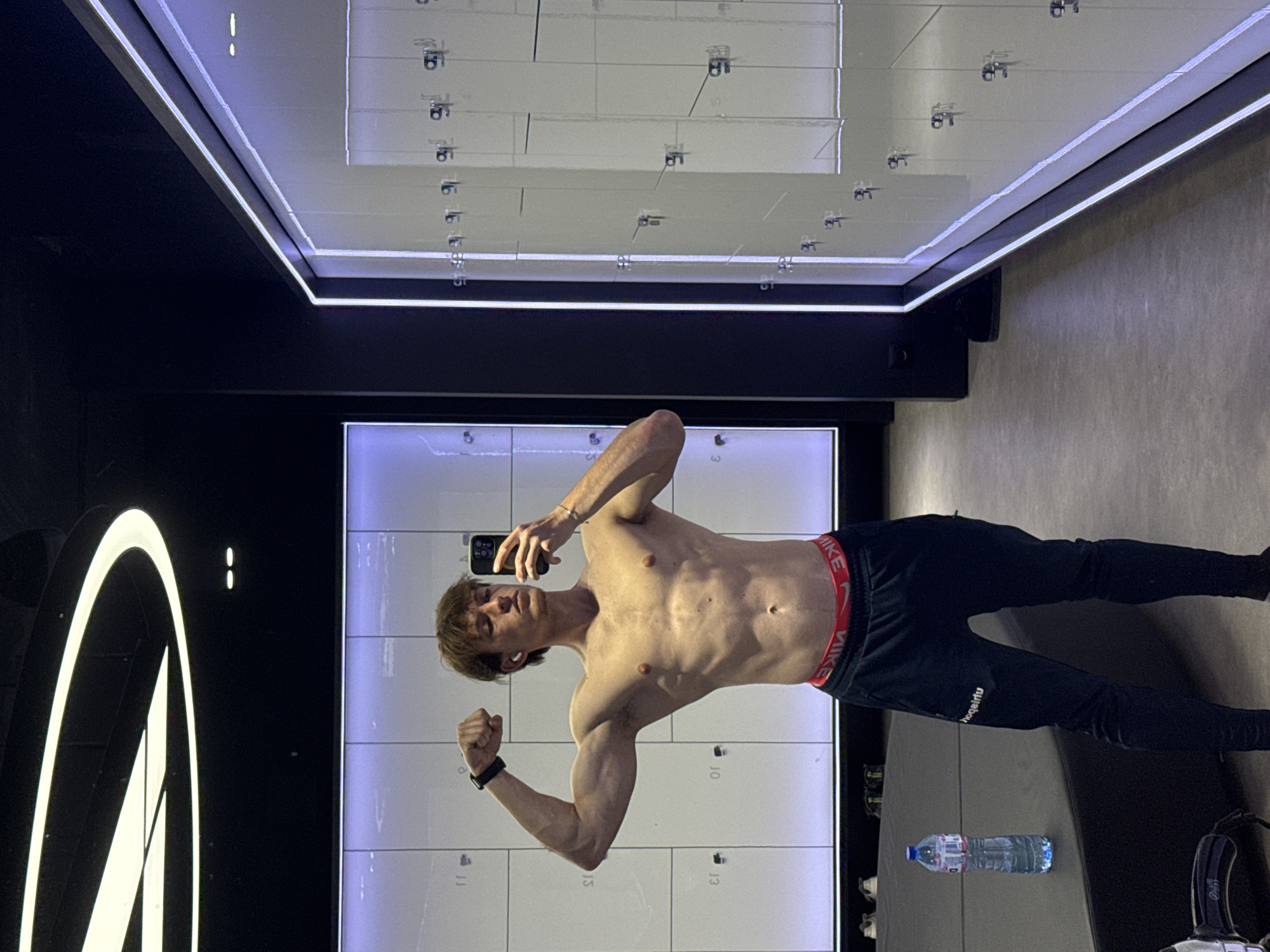 A shirtless young man with light brown hair is taking a mirror selfie inside what appears to be a modern gym changing room or locker area. He is posing with his left arm flexed, holding a smartphone in his right hand. He wears black athletic sweatpants with a red waistband bearing the "NIKE" logo, and the brand "hooqairtu" is visible (reversed due to the mirror) on his left pant leg. A black smartwatch or fitness tracker is on his left wrist.
The background features white lockers, some with numbers like "10," "11," and "13" visible. The room has dark walls and prominent LED strip lighting, including around a mirrored ceiling that reflects the lockers and lights. A large, illuminated white logo, possibly an abstract "M" or part of a brand name, is on the left wall.
On the floor, a clear plastic water bottle with a blue cap and label is visible, along with a pair of white shoes and what appears to be a black gym bag or equipment. The setting suggests a contemporary fitness center in Sofia, Bulgaria. The man's pose indicates he is documenting his physique, likely after a workout. The image is oriented with the man standing horizontally across the frame.