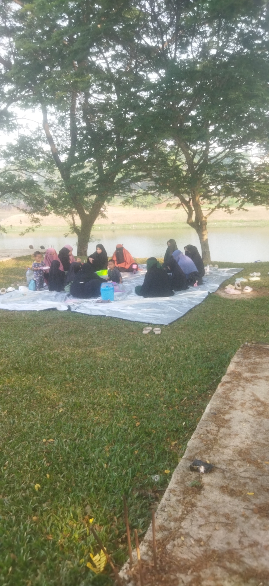 A group of individuals, including adults and children, are gathered on a white tarp spread on a grassy area near a body of water. Most of the adults are wearing hijabs and some are wearing face veils, indicating a possible Islamic gathering. They appear to be having a picnic or enjoying a social gathering outdoors. In the foreground, a concrete path with some debris is visible. The background shows trees and the water body, suggesting a park or recreational area. The lighting indicates it is daytime, possibly late afternoon given the warm tones. The overall scene depicts a relaxed outdoor activity in Depok, Indonesia.