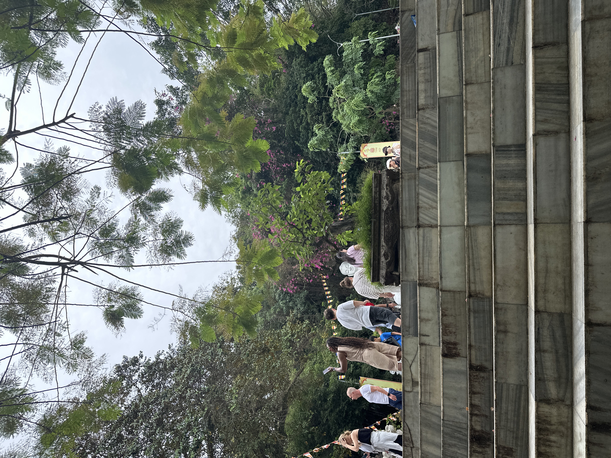The image depicts a scene at the Marble Mountains in Đà Nẵng, Vietnam. In the foreground, a wide set of marble stairs leads upwards. Several people are visible on and around the stairs. On the left, a woman in a white top and striped pants is looking up. Next to her, a man in a white t-shirt is standing. Further to the right, a woman in a light brown outfit is holding up a phone, appearing to take a picture. Behind her, a man in a white t-shirt and shorts is walking. A group of people in casual clothing are also visible on the stairs and in the background.
The background is dominated by lush greenery, with trees and flowering bushes, suggesting a natural landscape. Prayer flags are strung between trees, adding to the cultural context of the location. A sign with Vietnamese text is visible on the right side of the frame, and a white statue of Buddha's head is partially visible behind the people. The sky is overcast, indicating a cloudy day. The marble stairs are wide and appear to be made of large, grey slabs with natural veining.