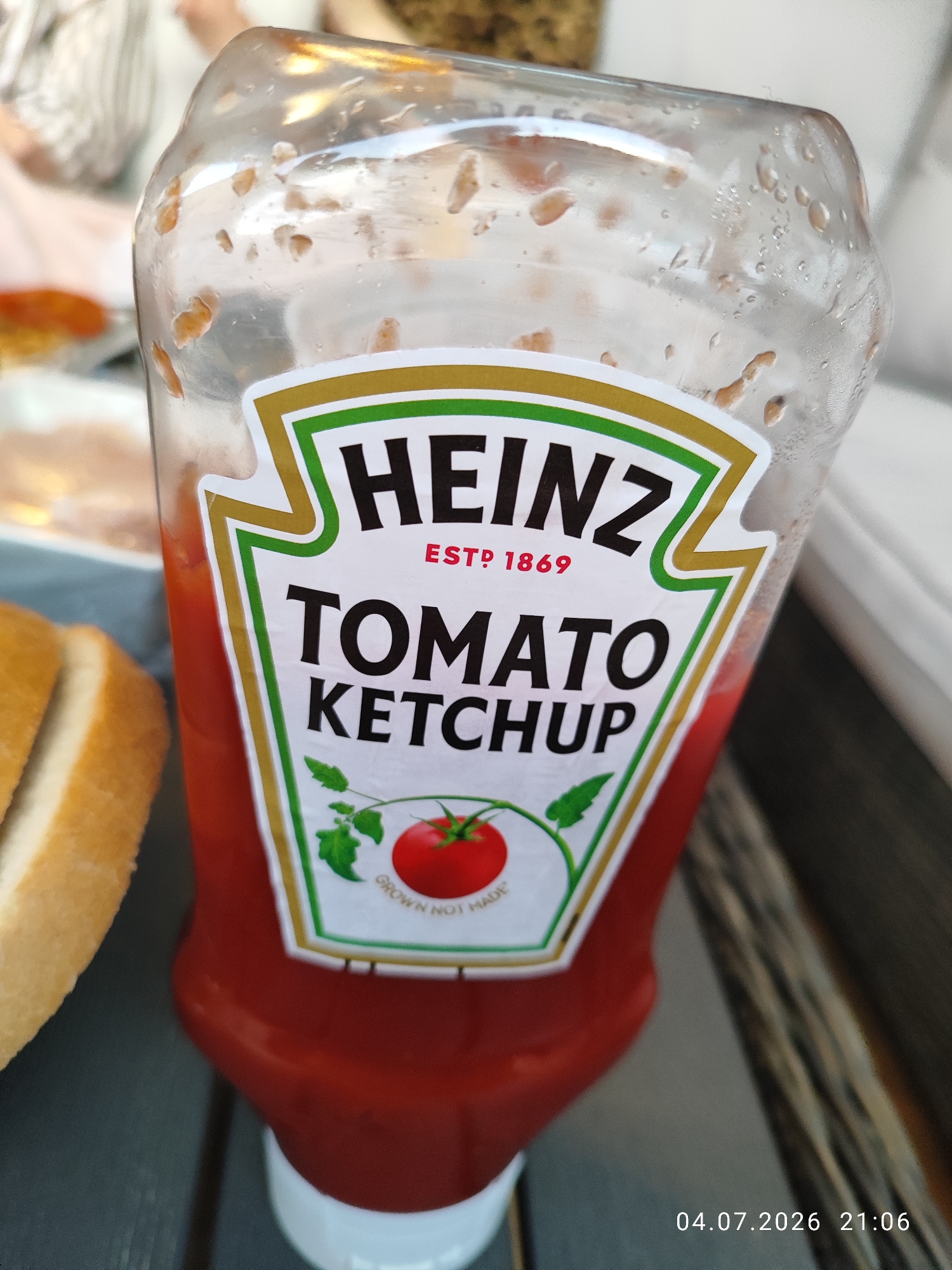 A close-up, eye-level, slightly angled shot shows a nearly empty plastic bottle of Heinz Tomato Ketchup. The bottle is mostly transparent, revealing smudges and water droplets on its interior and exterior surfaces. The label, a distinctive white shield with gold and green trim, is prominent. It features the "HEINZ" logo in large black text, followed by "EST. 1869" in smaller red text. Below that, "TOMATO KETCHUP" is printed in bold black lettering. A graphic of a tomato plant with a single red tomato is also visible on the label. The ketchup inside the bottle is a deep red color. The bottle's white cap is at the bottom of the frame. To the left of the bottle, the edge of a slice of bread or bun is visible. The background is blurred, with hints of people and possibly food items. The scene appears to be outdoors, with a wooden surface in the foreground and a suggestion of a table. A date stamp "04.07.2026 21:06" is superimposed in the bottom right corner. The image does not provide any specific landmarks or visual cues to indicate a location within the city of Wellingborough, United Kingdom.