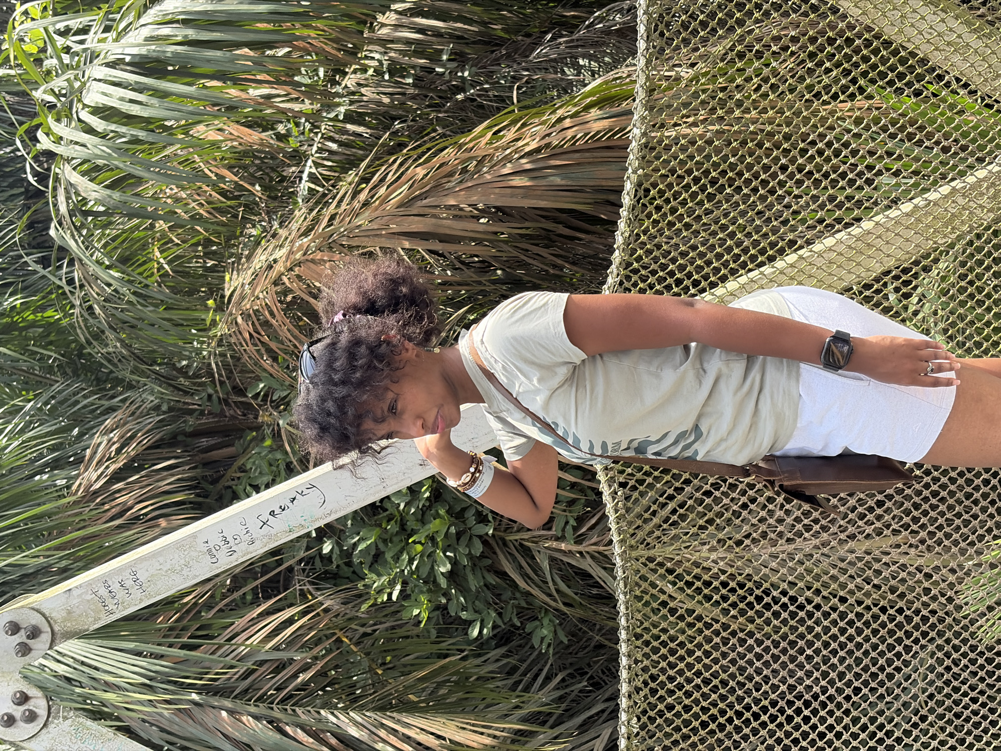 A woman with curly black hair stands on a suspended net walkway, likely part of a canopy walk, in Lekki, Nigeria. She is wearing a light-colored t-shirt and white shorts. Her left arm rests on a white metal beam of the walkway, which is covered in graffiti. A brown shoulder bag hangs from her right shoulder. She is wearing a watch and bracelets on her wrists. Green foliage, including palm fronds, surrounds the walkway. The location is likely the Lekki Conservation Centre.