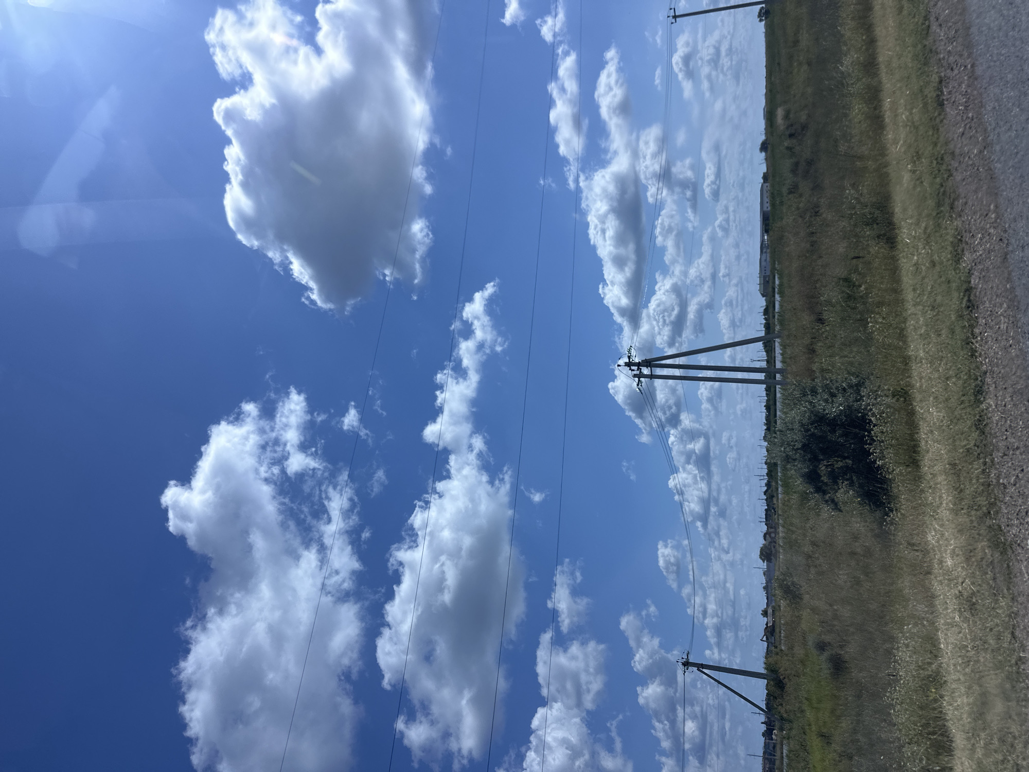 The image depicts a rural landscape under a bright blue sky dotted with fluffy white clouds. In the foreground, dry, tall grass and some scrubby bushes cover the ground, with a gravel path visible at the bottom edge. Several power poles with electrical wires stretch across the frame, some in the middle ground and others more distant. In the background, there's a low horizon line with a few scattered buildings and structures, suggesting a small settlement or industrial area. The overall impression is one of a sunny, open, and somewhat sparse environment. The location is identified as Peresyp, Russia.