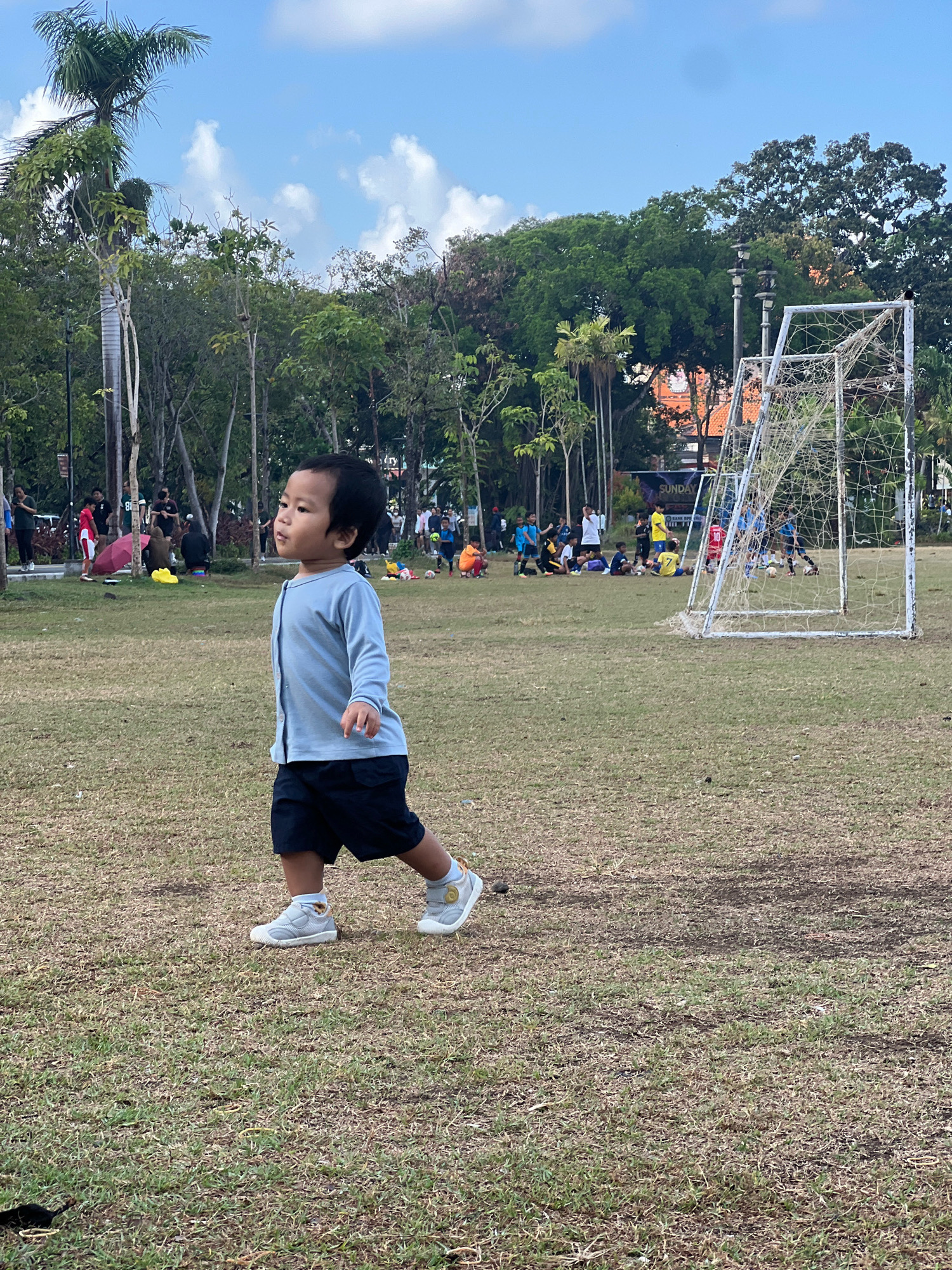 A young child is walking in a grassy field. The child is wearing a light blue long-sleeved shirt and dark shorts. They are also wearing white sneakers with grey accents. The child is looking to the left of the frame. In the background, there are trees, a soccer goal, and other people playing soccer. The scene appears to be in a park.
The image was taken at Lapangan Banteng in Denpasar, Indonesia. This is a large, open field commonly used for sports and recreation. The presence of a soccer goal suggests that the area is frequently used for soccer games and practices. The surrounding trees and lush greenery indicate a tropical environment, consistent with Denpasar's climate. The sky is partly cloudy with patches of blue sky visible. The lighting suggests it is daytime, likely late afternoon given the angle of the sun and the soft shadows.