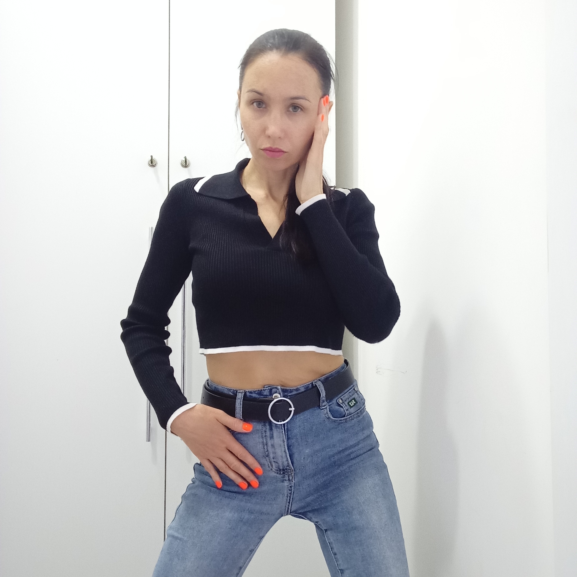 A woman stands indoors, posing in front of white cabinets. She is wearing a black long-sleeved crop top with white trim, high-waisted light blue jeans with a black belt, and has bright orange nail polish. She is touching her face with her left hand, and her right hand rests on her hip. She appears to be in a room with a simple, white interior. There is no visible evidence to confirm or deny that the location is in the city of Izhevsk, Russia.