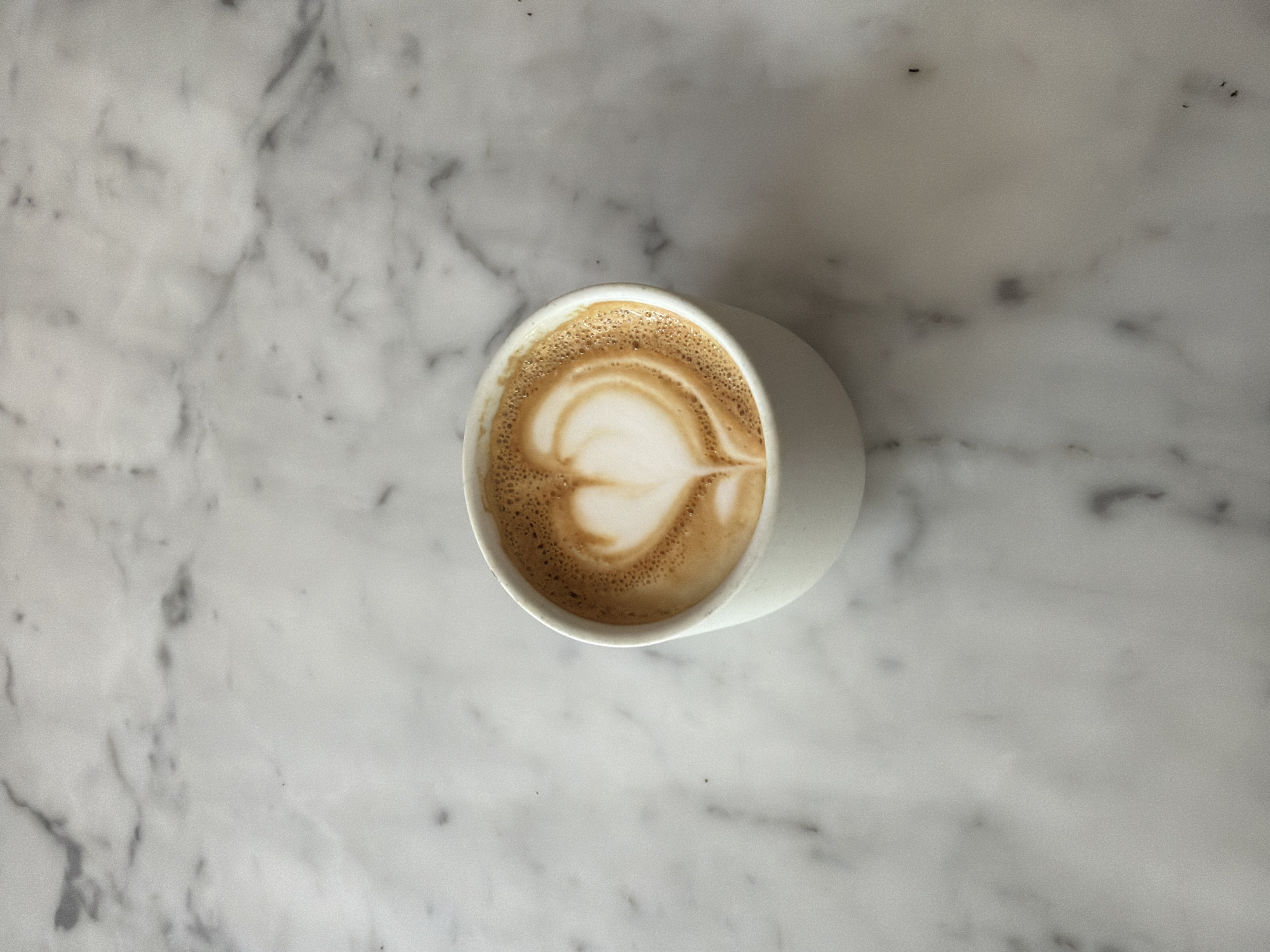 This overhead shot captures a single white ceramic cup filled with coffee, artfully prepared with latte art in the shape of a heart. The coffee, likely a latte or cappuccino, features a rich brown espresso base topped with creamy white foamed milk, forming a distinct heart pattern. Small bubbles are visible on the surface of the foam, indicating freshness.
The cup rests on a clean, polished white marble surface, characterized by subtle grey veining that adds texture and depth to the background. The marble table extends beyond the edges of the frame, providing a minimalist and elegant setting. The lighting is soft and even, suggesting a bright, natural illumination, likely indoors during daytime. There are no harsh shadows, contributing to the clean aesthetic.
The image focuses solely on the coffee and its immediate surroundings, creating a sense of calm and simplicity. There are no people visible, and no specific activities other than the implied preparation and readiness for consumption of the coffee. No text is visible in the image. The overall impression is one of modern style and attention to detail, typical of contemporary cafe culture, potentially in Los Angeles.