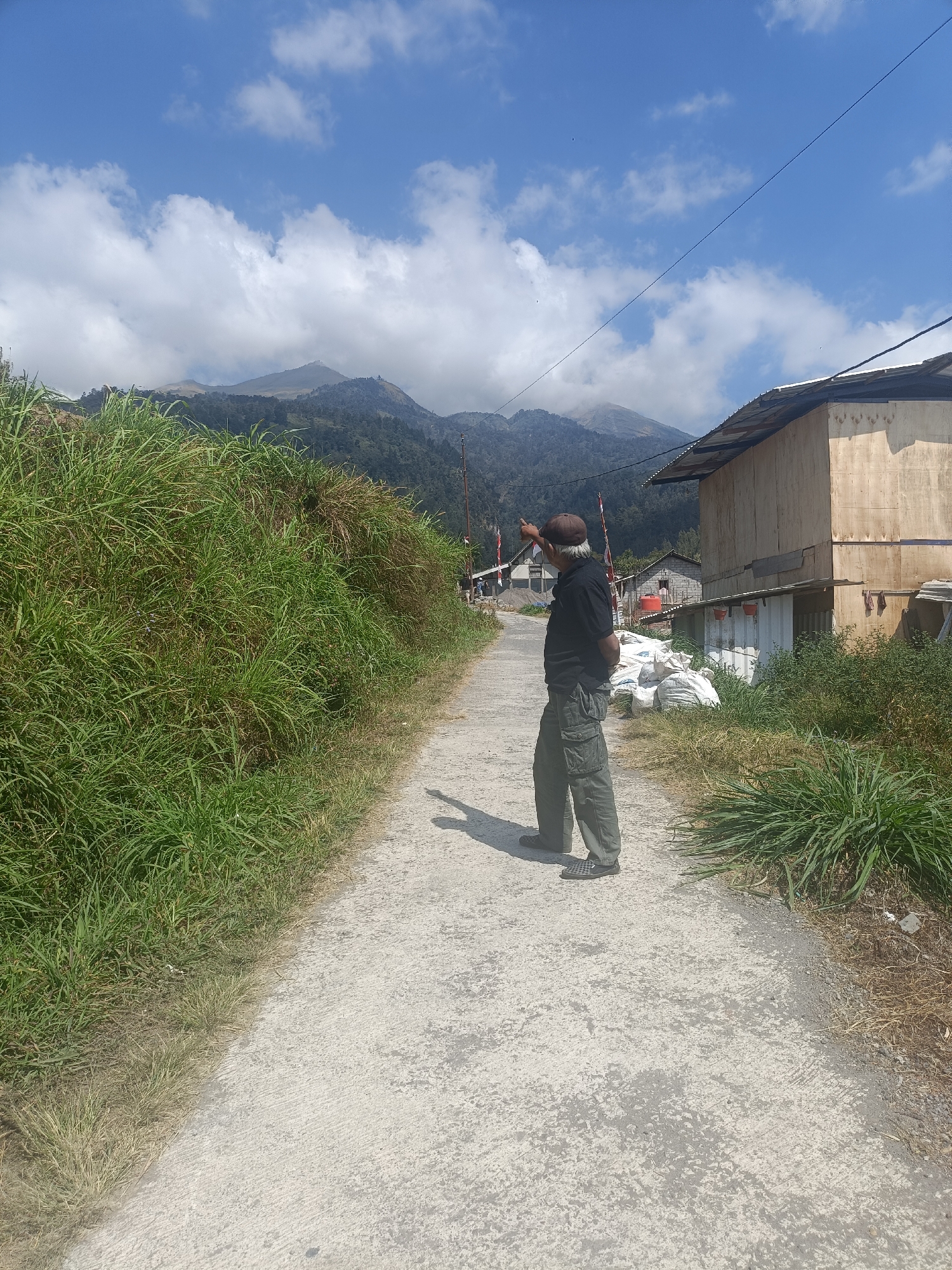 The image depicts an outdoor scene in the village of Kopeng, Indonesia, likely in a rural or mountainous area. In the foreground, a man wearing a dark shirt, cargo pants, and a brown cap stands on a paved pathway, pointing towards the distant mountains. He is seen from the back, with his left arm raised. Lush green grass and foliage border the pathway on the left, while on the right, there is a simple wooden building and several large white sacks, possibly containing goods. In the background, a majestic mountain range rises under a bright blue sky dotted with white clouds. The overall atmosphere suggests a scenic and possibly agricultural setting.