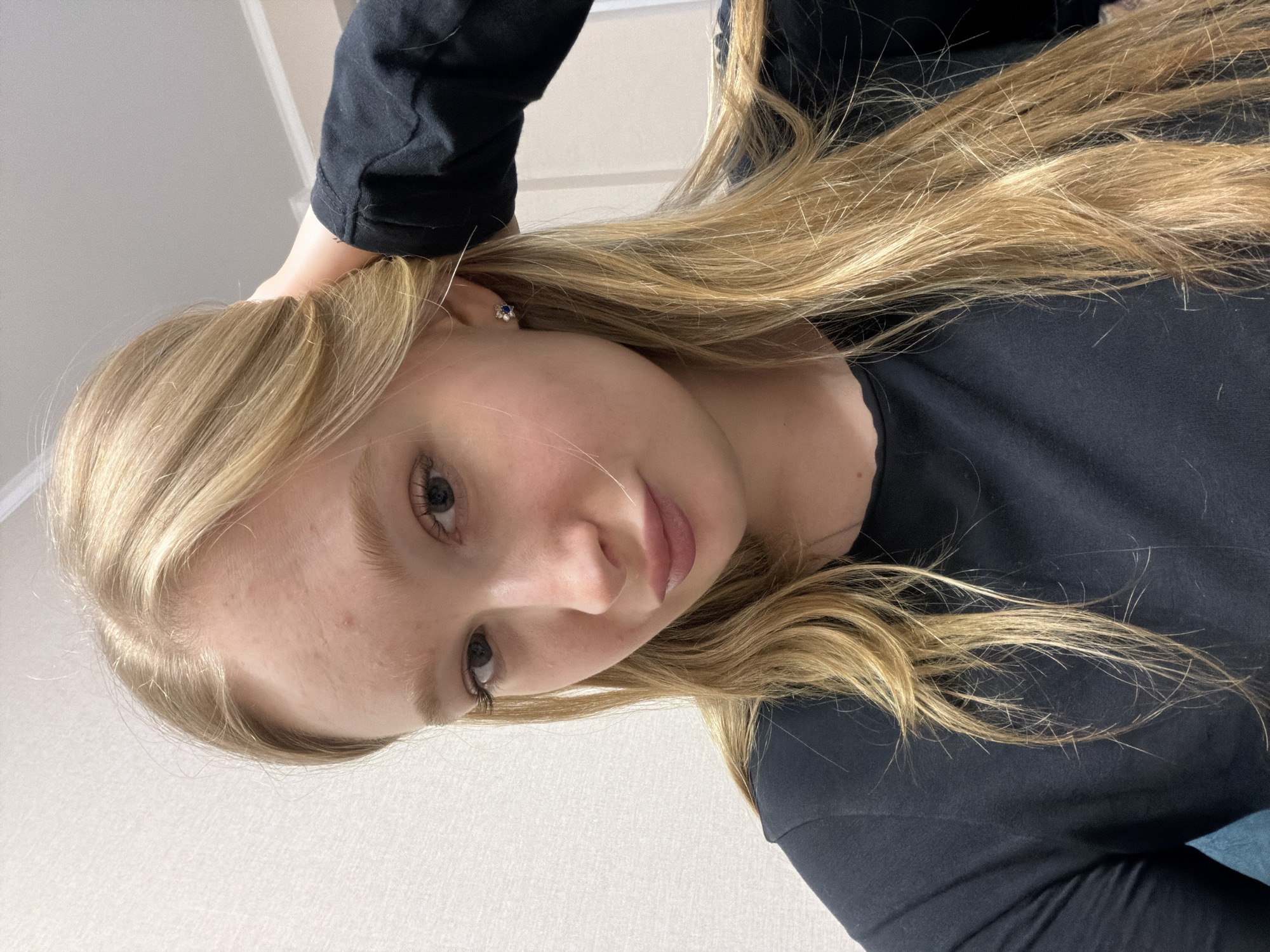 A young woman with blonde hair is shown in a close-up, tilted head-and-shoulders portrait. She is wearing a black long-sleeved shirt and a stud earring is visible in her left ear. Her hand is near her head. The background appears to be a plain, light-colored wall. There is no information available to determine the location of this image as Засечное, Russia.