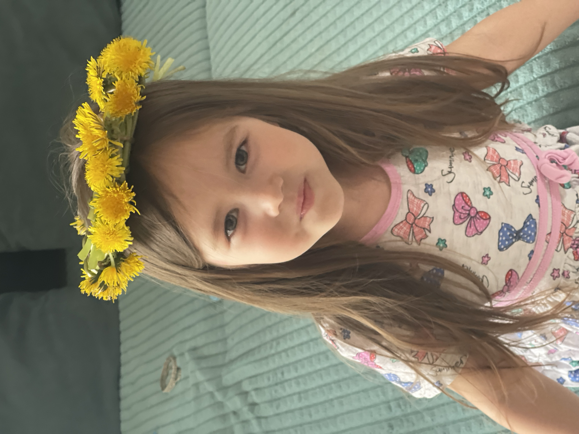 A young girl with long brown hair wears a wreath of yellow dandelions on her head. She is wearing a light-colored top with a pattern of bows and other designs. The background is a textured, turquoise-colored surface. The image was taken in Verkh-Katunskoye, Russia.