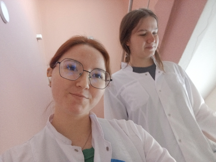 The scene shows two young females indoors. The specific location within Chaikovsky, Russia, cannot be determined from the image. The subject in the foreground has reddish-brown hair, wears large, dark-framed glasses, a white earbud in her left ear, a green shirt, and a white lab coat. She is looking at the camera with a subtle smile. The second subject, positioned behind and to the right, has long brown hair, wears a dark shirt, and a white lab coat, with her gaze directed slightly away from the camera. Both individuals are attired in white lab coats. The immediate environment consists of plain, light pink walls. A circular white light fixture and a smaller white rectangular fixture are present on the wall to the left of the foreground subject. Illumination is bright and uniform.