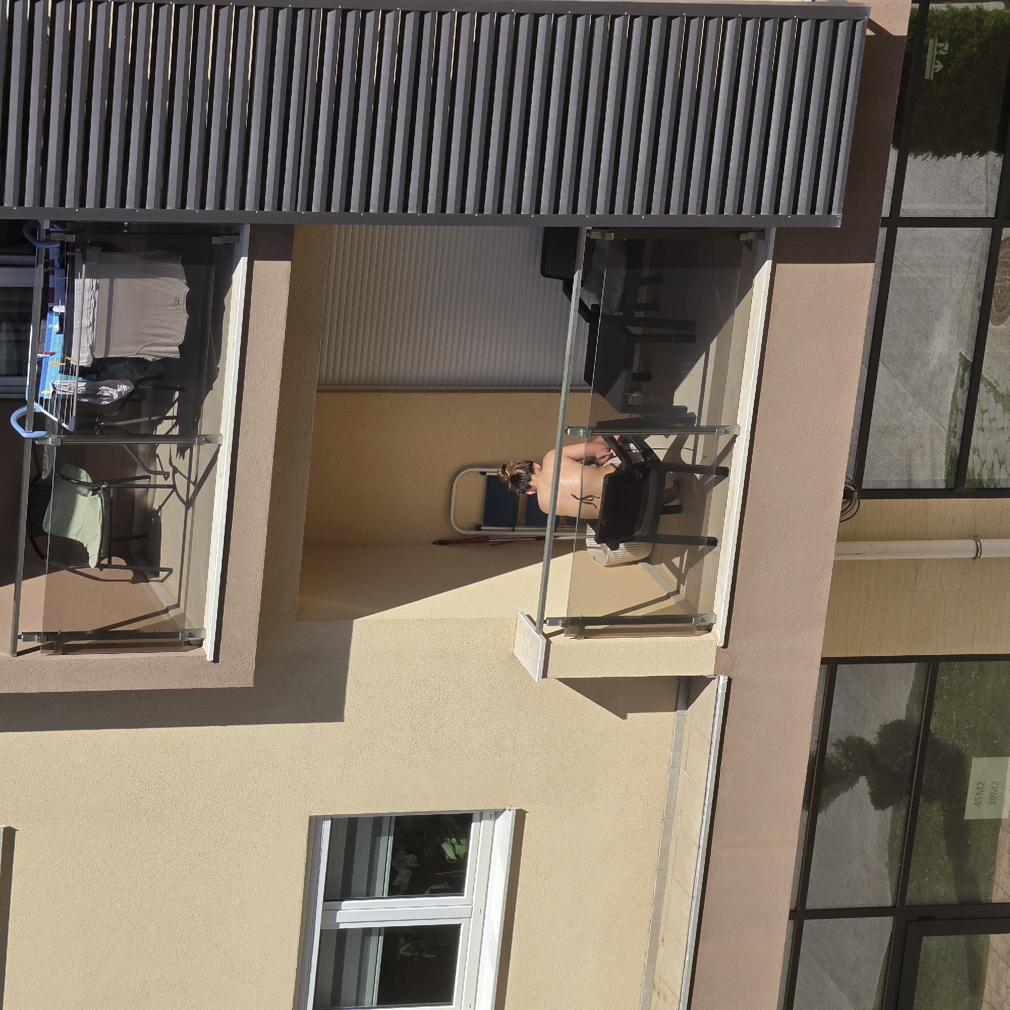 A top-down view shows a portion of a building with balconies in Podgorica, Montenegro. A woman with her hair in a bun, is sunbathing on one of the balconies, sitting in a black chair. Clothes are drying on a clothes rack on the left balcony. A table and chairs are also on the balcony. A ladder stands behind her, and the balcony has a glass railing. A building window is visible below the balcony. The surrounding environment includes architectural details and greenery.