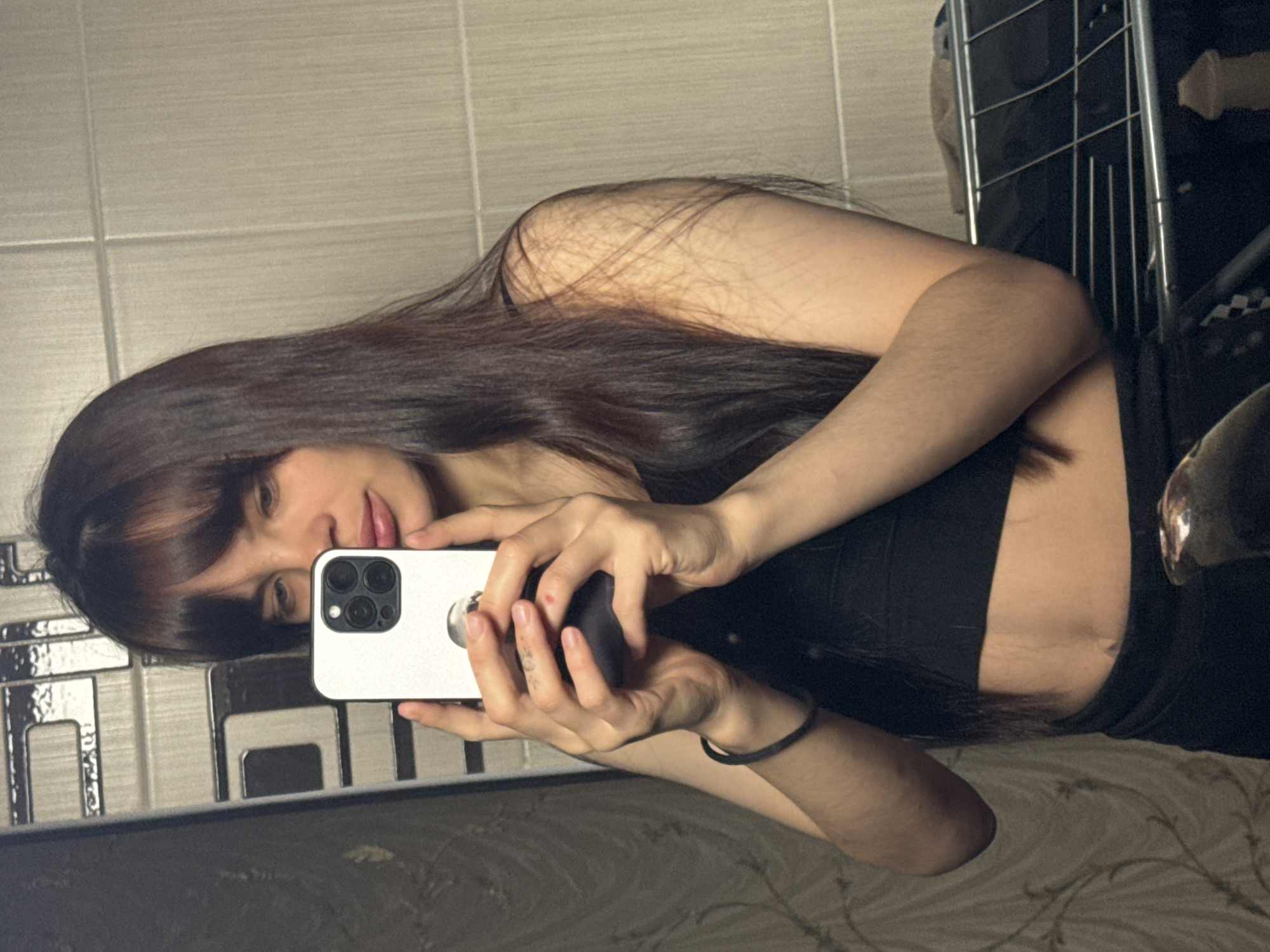 The image shows a young woman taking a mirror selfie in an indoor setting. She has long, dark brown hair with bangs and fair skin, and is wearing a black top that reveals a portion of her midriff. Her facial expression is subtle, with a slight smile and her eyes directed towards the phone.
She holds a white smartphone, likely an iPhone Pro model given its distinct multi-lens camera system, in her right hand. Her left hand is partially visible, holding a dark, indeterminate object, possibly a phone accessory. A black hair tie is worn on her left wrist. On her right side, a silver-colored buckle is visible on what appears to be a belt or part of her clothing.
The background features a light-colored tiled wall with horizontal grout lines. To her right, a dark metal rack or shelving unit is present. Below, a patterned fabric, possibly bedding or a blanket, is visible. The lighting suggests an indoor environment. No visible text appears in the image.