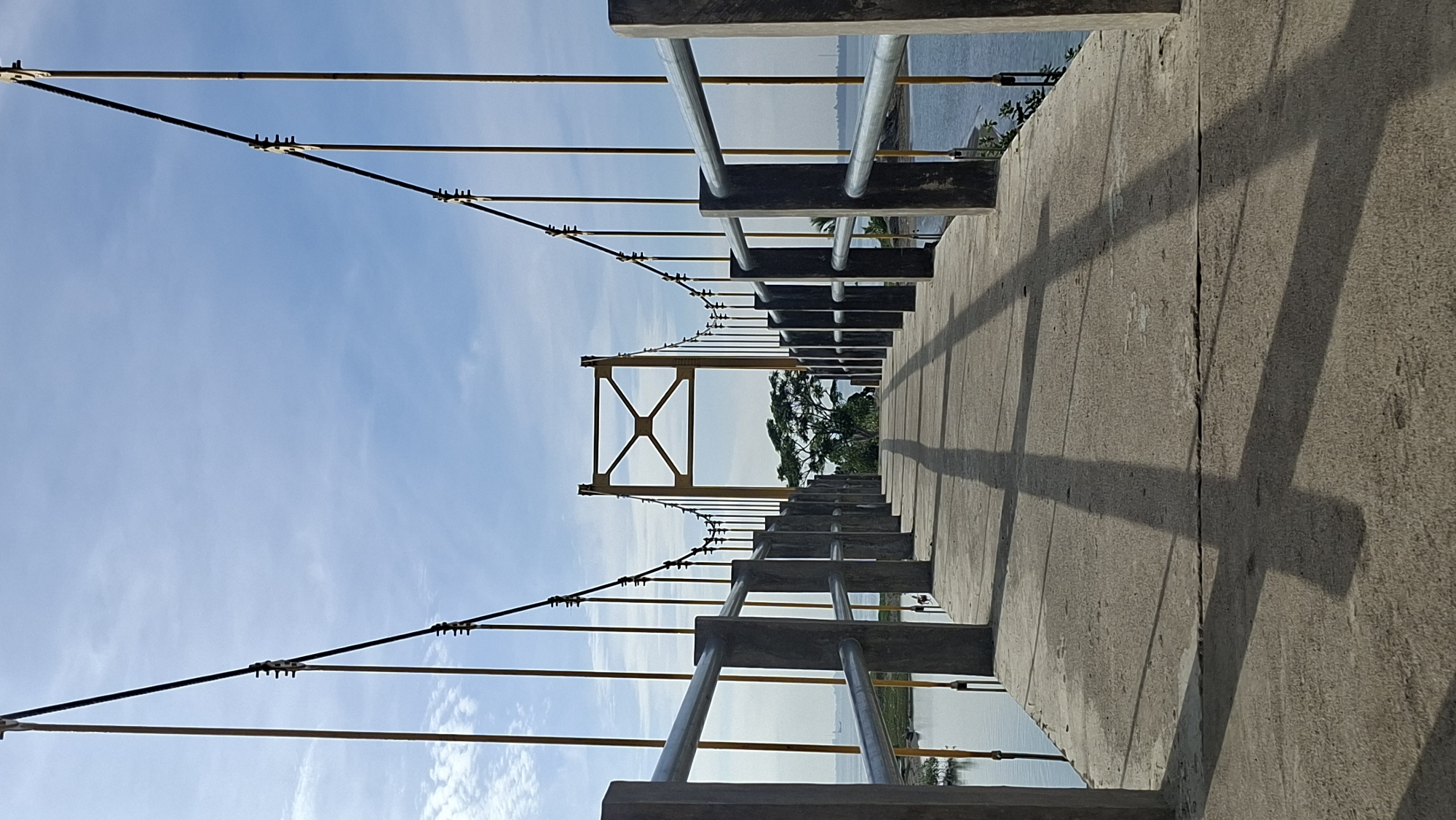 A low-angle, full-shot image captures a concrete pedestrian suspension bridge on a clear day, with a bright blue sky dotted with wispy white clouds above. The bridge, leading into the distance, features sturdy concrete pillars supporting horizontal metal railings on either side. Parallel to the railings, slender yellow poles rise, connected by dark cables that form an arc across the sky, suspending the bridge. The roadway of the bridge is concrete, showing faint lines from its construction. Long, distinct shadows of the railings and vertical supports stretch across the bridge, indicating the sun is positioned to the left and slightly behind the viewer.
In the middle distance, a bright yellow, A-frame suspension tower stands, supporting the main cables of the bridge. Beyond the tower, a single, prominent tree with green foliage stands on a distant shore. The water on both sides of the bridge appears calm, with a slight shimmer reflecting the sky. The environment suggests a coastal or riverside location, with a hint of the horizon visible on the left, where a small ship or structure can be seen in the far distance, and a sliver of beach on the right. This bridge is likely located in Lhokseumawe, Indonesia, a coastal city known for its ports and bridges over waterways. No people are visible in the frame.