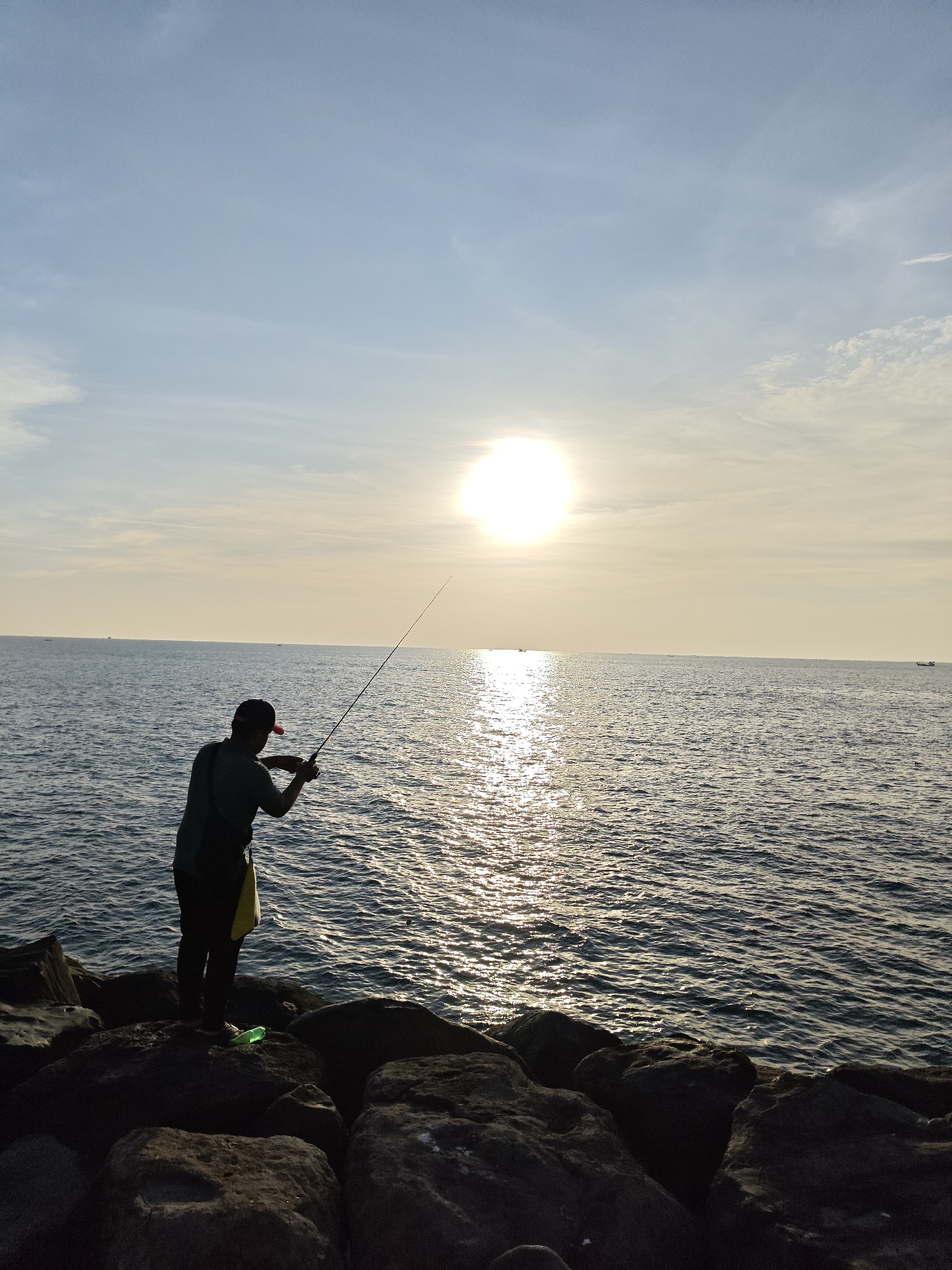 A person is fishing at the seafront in Lhokseumawe, Indonesia. The individual is positioned on dark-colored rocks with a fishing rod extended over the water. A green water bottle is on the rocks. The background includes a body of water with the sun reflecting off the surface, and a sky with some cloud cover. Several distant boats are visible on the horizon.