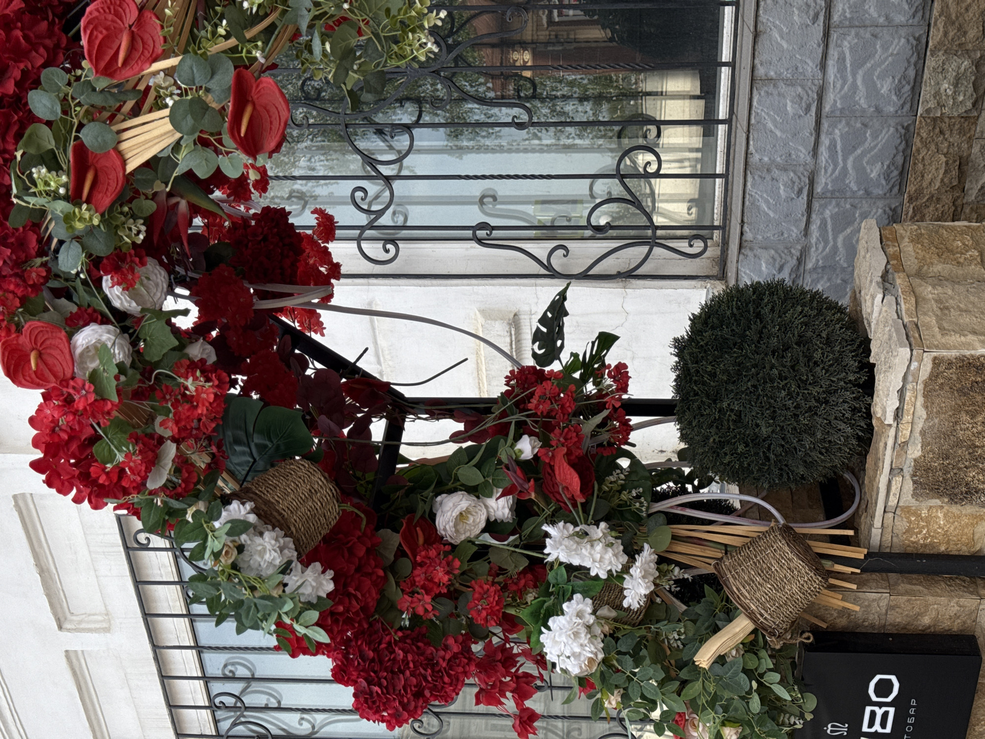 The image shows a floral arrangement and a trimmed bush outside a building. The arrangement is primarily composed of red and white flowers, green foliage, and woven baskets. A metal grate or railing is visible behind the arrangement. The trimmed bush is round and dark green. The building's exterior features a combination of light and dark stone or brickwork. The location is Sarapul, Russia.