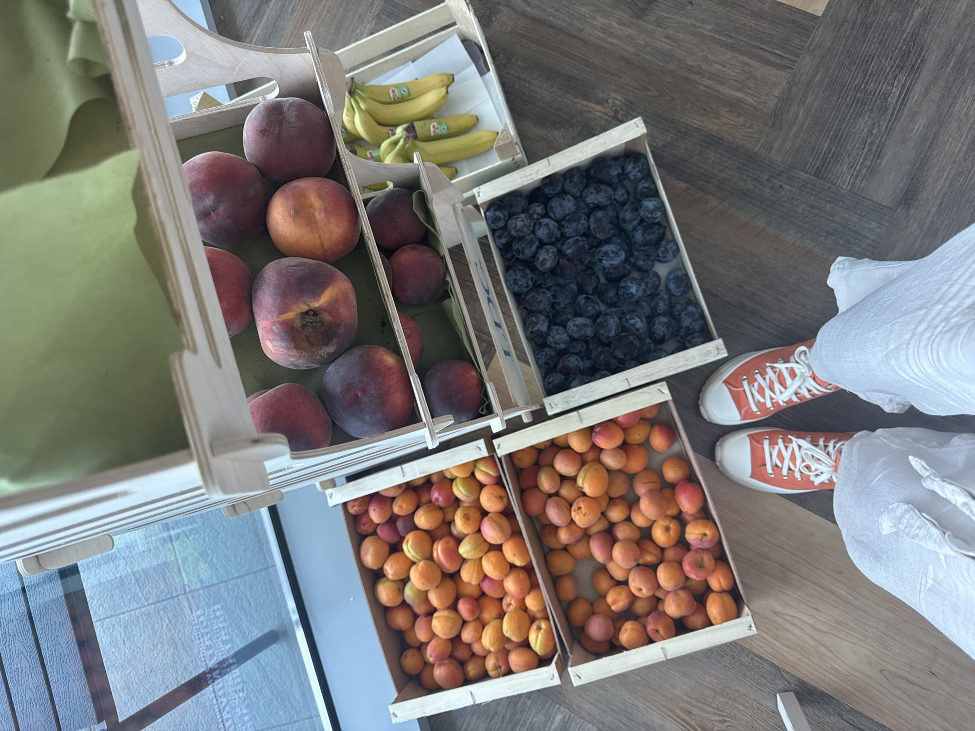 The image shows a fruit stand with two shelves filled with various fruits. On the top shelf, there are several peaches. The middle shelf contains two crates of apricots and a crate of bananas. The bottom shelf has a crate filled with plums. In the foreground, a person wearing white pants and orange sneakers is visible, standing in front of the fruit stand. The background appears to be a street scene through a window, suggesting an outdoor or semi-outdoor location. The overall impression is a display of fresh produce, possibly in a market or a shop. The presence of a fruit stand and the general setting suggest a commercial establishment. The image does not contain any specific visual cues that would allow for precise identification of the location in Sofia, Bulgaria.