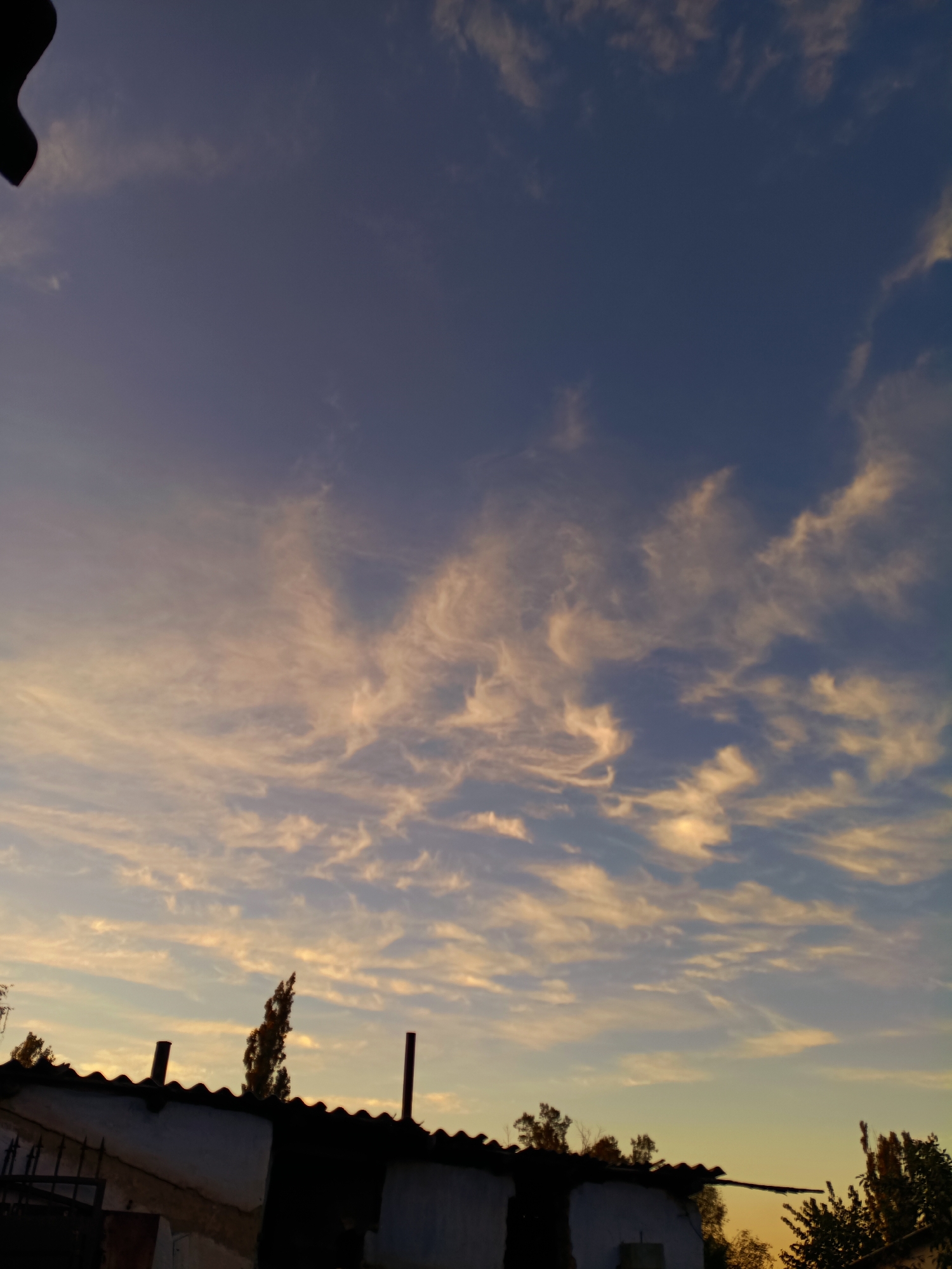 This is a vertical, low-angle shot of the sky at dusk over Tash-Dobo, Kyrgyzstan. Wispy, elongated clouds, tinged with a pale orange and yellow from the setting sun, are scattered across a deep blue sky. Silhouetted against the colorful sky are the corrugated roofs of several low-lying buildings. The roofs have a scalloped edge. Two dark, cylindrical chimney pipes protrude from one of the roofs. A tall, thin tree with autumnal foliage stands between the chimney pipes. The foreground is dominated by the dark, angular shapes of the rooftops and the walls of the buildings, rendered in silhouette against the brighter sky. A dark, curved object, possibly part of a larger structure or an overhanging tree branch, is visible in the upper left corner of the frame. No people are visible in the image.