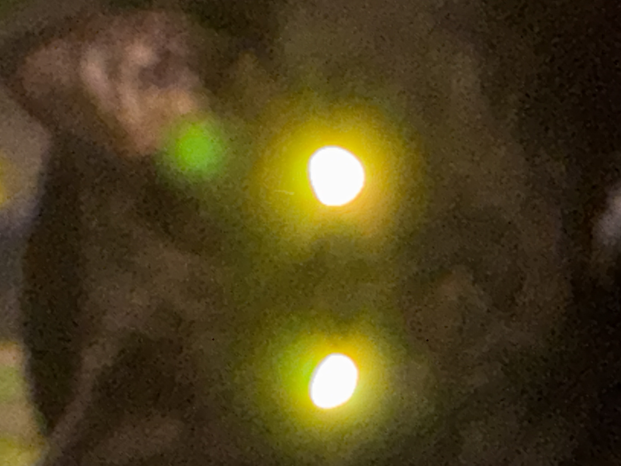 A blurry, low-resolution image shows two distinct orbs of bright yellow light against a darker, mottled background. At the top left of the frame, a less defined, greenish area is visible. The overall scene appears to be photographed in low-light conditions. The location is not discernible from the image. There are no identifiable people, specific subjects, objects (beyond the lights), actions, or interactions visible in the frame. The environment is vague and indistinct.