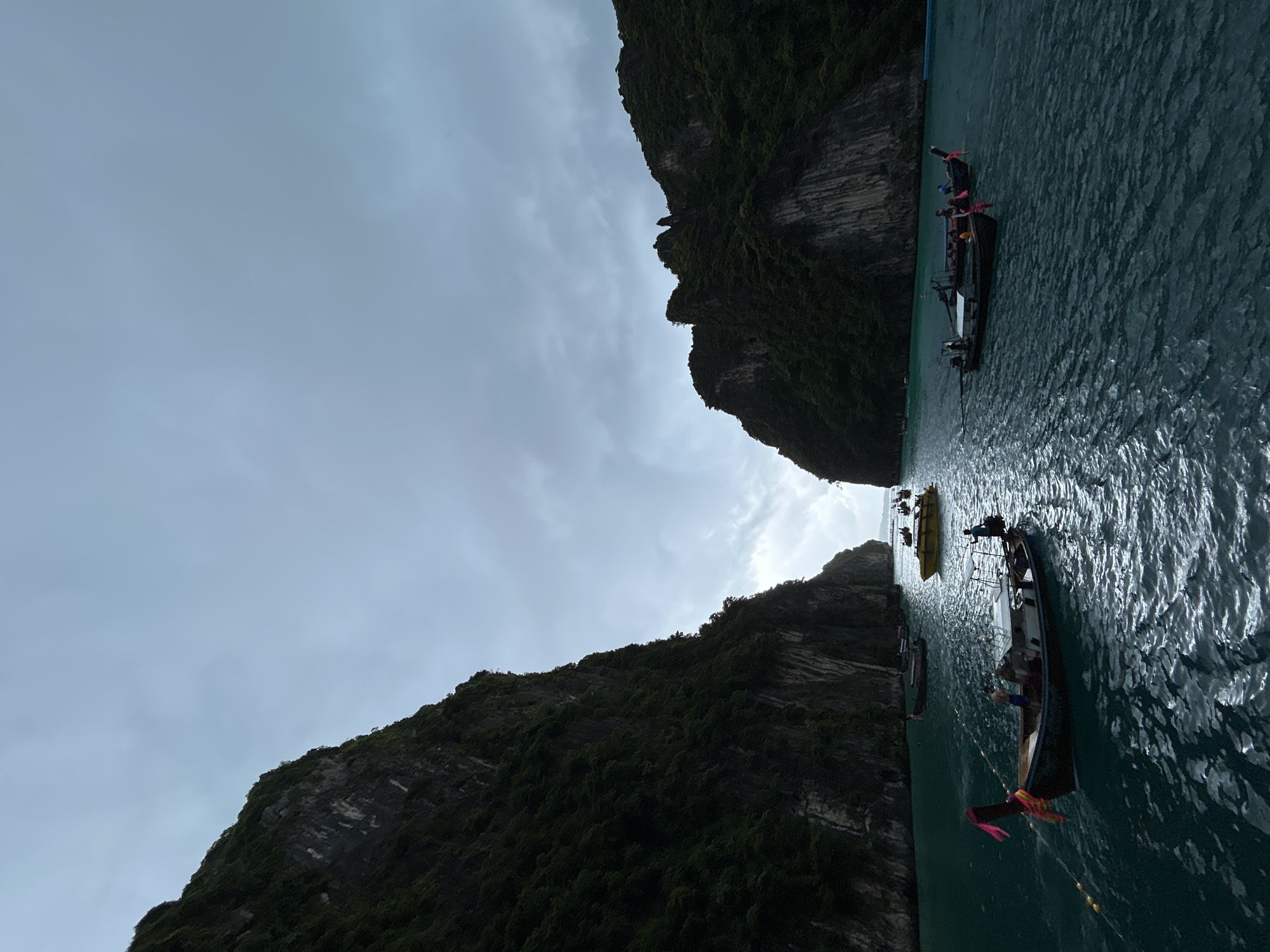 The image depicts a serene bay in Ao Nang, Thailand, flanked by towering, verdant limestone cliffs. The water is a deep, tranquil turquoise, with gentle ripples reflecting the overcast sky. Several longtail boats, characteristic of Thai maritime transport, are scattered across the bay. One boat in the foreground, adorned with colorful fabric streamers, has two individuals visible onboard. Another similar boat is moored nearby. Further in the distance, a yellow speedboat navigates the waters, accompanied by other longtail boats. The overall atmosphere is one of peaceful natural beauty, with the dramatic cliffs framing the tranquil scene.