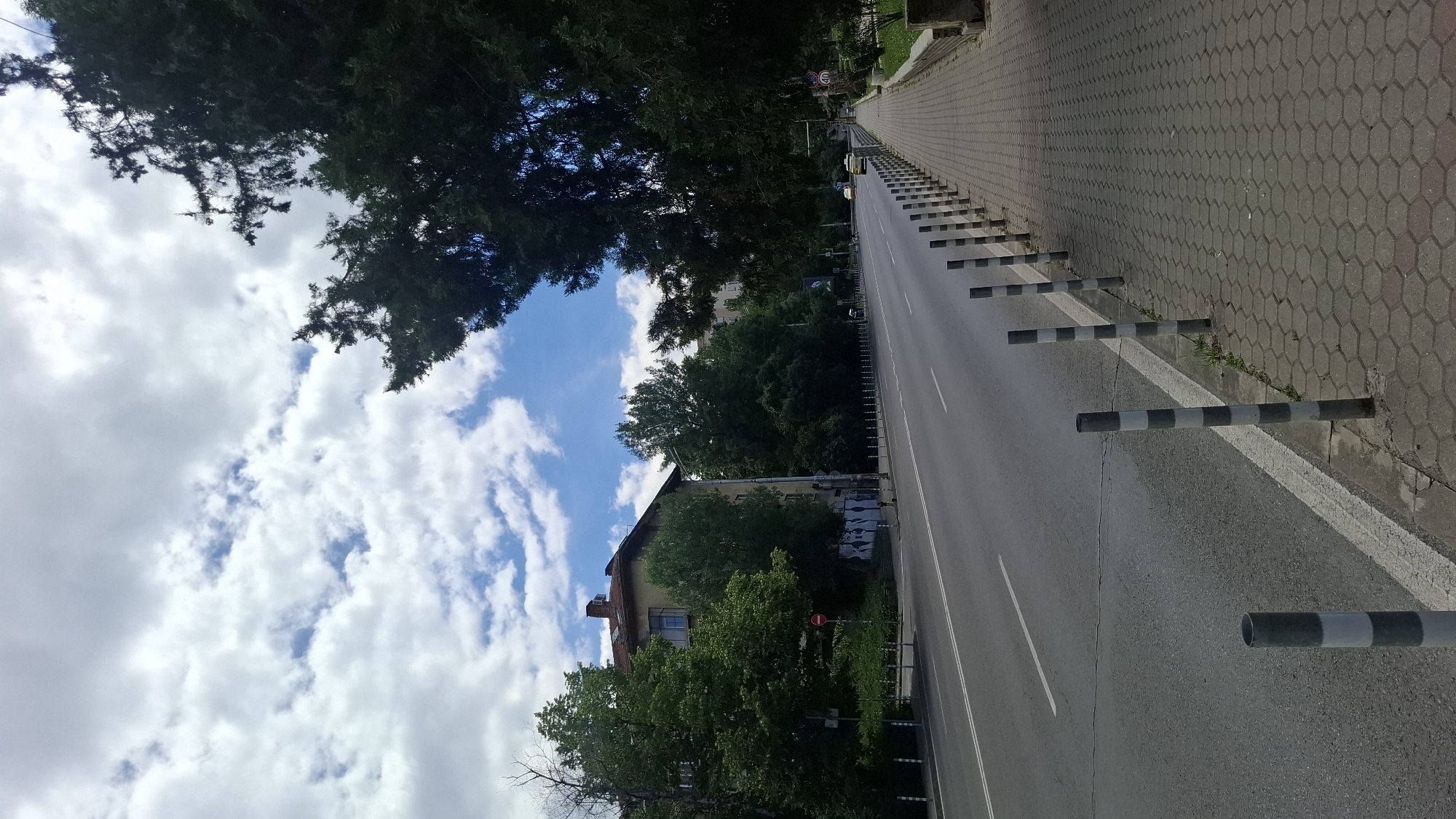 This is a medium shot of a street in Sofia, Bulgaria, on a cloudy day. The street is asphalt and appears to be a main thoroughfare with multiple lanes. A paved sidewalk runs along the right side of the street, separated by a row of bollards. The sidewalk is made of hexagonal grey pavers. Trees line the left side of the street, behind the sidewalk. A building with a red-tiled roof is visible on the left, partially obscured by trees. The sky is filled with white and grey clouds. The overall scene depicts a typical urban streetscape.