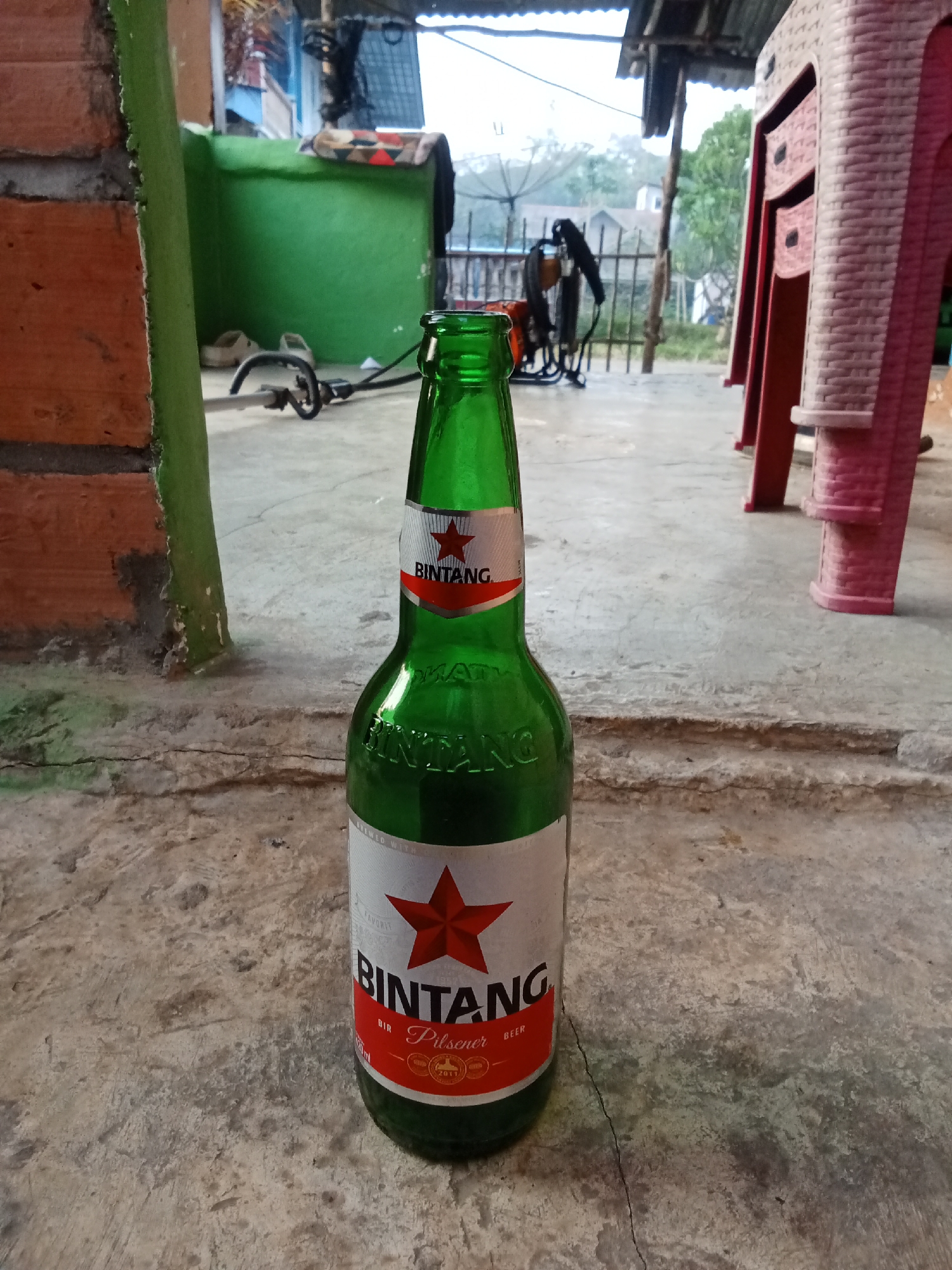 A Bintang beer bottle stands upright on a concrete surface in Tebing Bulang, Indonesia. The bottle is green glass with a white label featuring a red star and the brand name "BINTANG" in black lettering. The label also indicates "Pilsener" and "BEER." In the background, a green wall, a brick pillar, and red plastic chairs are visible, along with some vegetation and a structure with a corrugated metal roof. The scene appears to be outdoors, possibly a yard or patio area.
