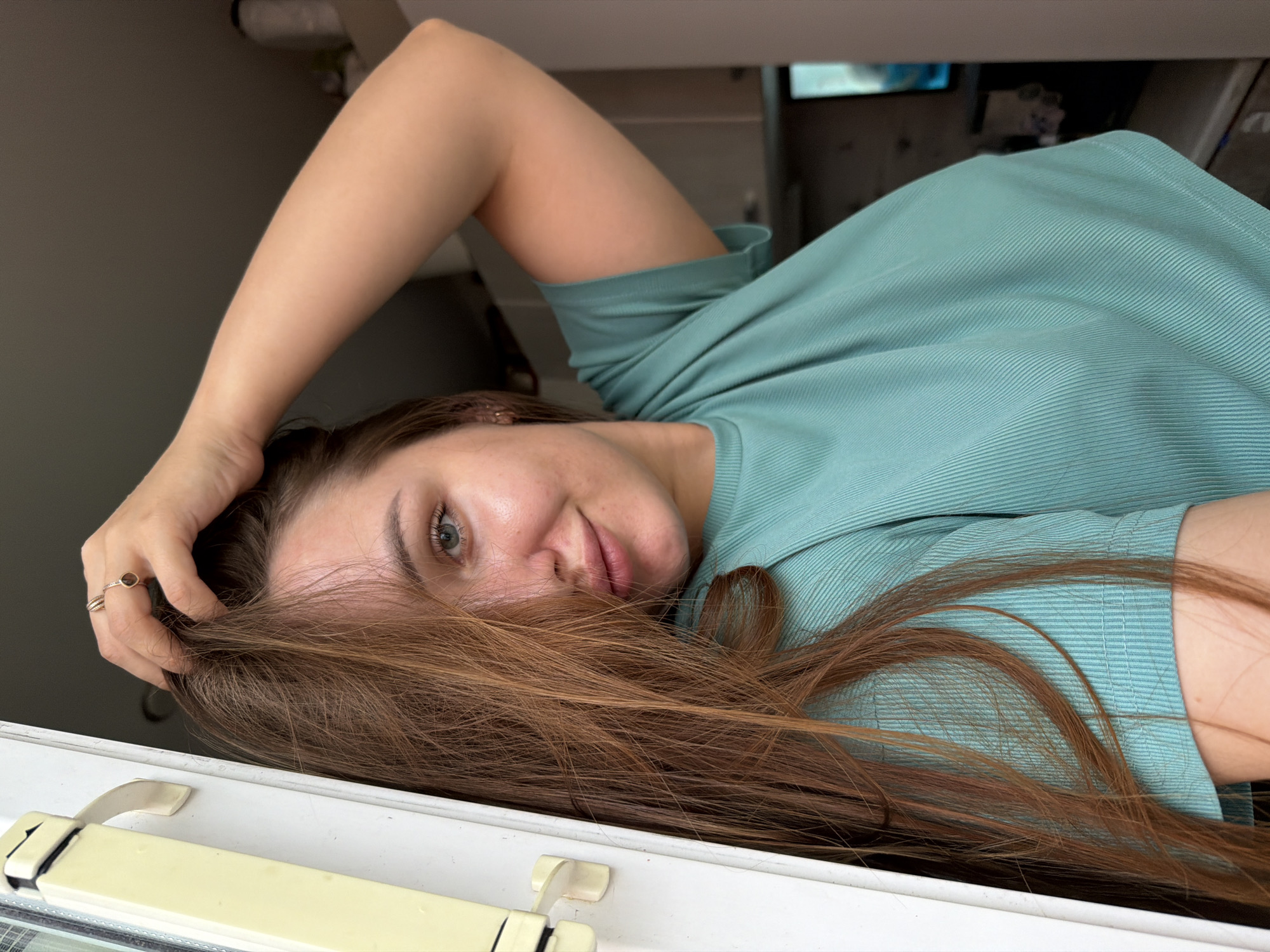 An eye-level, medium-shot photograph captures a young woman with long, brown hair, partially obscuring her face as she touches the top of her head with her right hand. She is wearing a turquoise, ribbed t-shirt. The woman is positioned in front of a light-colored door frame and a slightly out-of-focus background that appears to be an indoor setting with kitchen cabinets. The lighting suggests a bright, natural light source from the left, possibly from a window. The image does not provide any information that would indicate it was taken in the city of Chaikovsky, Russia.