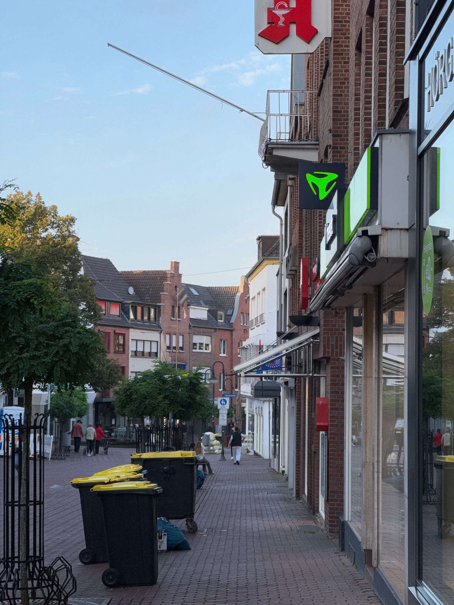 The image shows a street scene in Cleves, Germany. On the right side of the image, a brick building houses several shops, including a pharmacy with a red cross logo. A bright green logo of a three-pronged shape is also visible on the building. Storefront windows reflect the opposite side of the street. The street is paved with red bricks, and on the left side, there are trees, a metal fence, and several large black trash bins with yellow lids. A few people are walking on the sidewalk, and in the distance, more buildings are visible. The sky is blue with a few scattered clouds, and the lighting suggests it is either early morning or late afternoon.