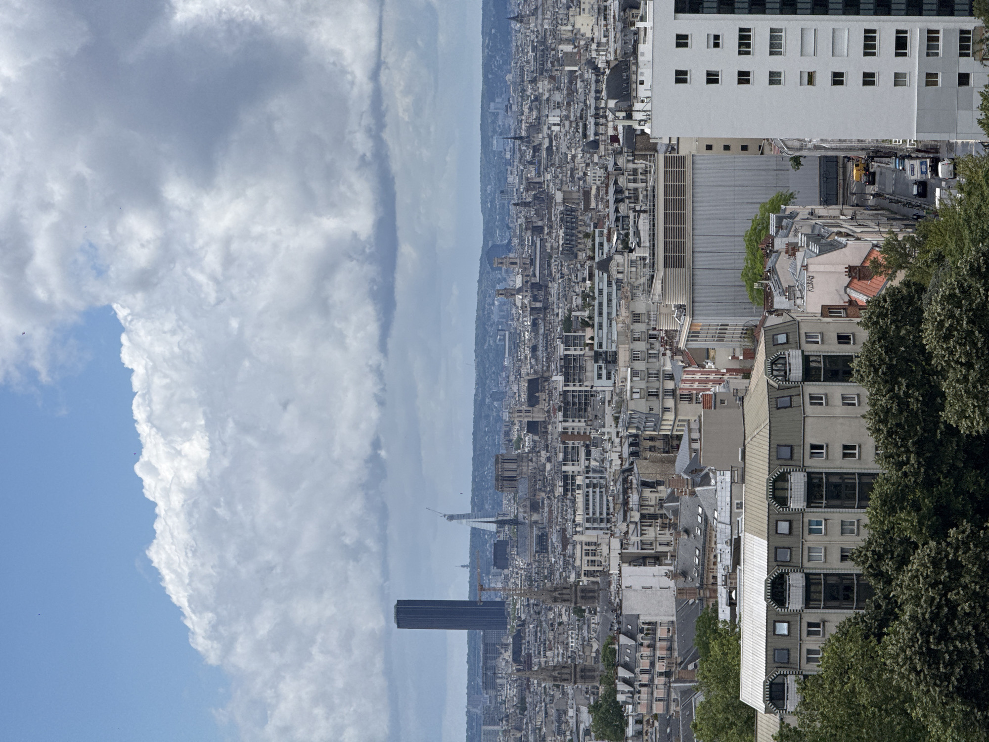 This detailed image depicts a panoramic view of Paris, France, observed from a high vantage point in the southeastern part of the city, likely from a tall residential building in the 13th arrondissement, looking generally northwest across the urban landscape.
The upper portion of the image is dominated by a dynamic sky filled with large, volumetric cumuliform clouds, varying from bright white to shades of grey, indicating an overcast but bright day. Patches of clear blue sky are visible, particularly in the upper left quadrant. Small, dark specks, possibly birds or distant aircraft, are discernible against the sky and clouds.
The horizon is a hazy line of distant hills or elevated terrain. The mid-ground and distant cityscape reveal numerous iconic Parisian landmarks. Prominently on the left, the dark, monolithic Tour Montparnasse stands tall. To its right, the distinctive, modern Tour Triangle is visible under construction, characterized by its pyramidal form and scaffolding with a crane. Towards the center of the image, the Notre Dame Cathedral is identifiable, undergoing restoration with visible scaffolding and a large crane around its structure. Numerous other church spires, domes, and the dense, uniform architecture characteristic of Parisian buildings extend across the vast urban sprawl.
The foreground features a mix of urban architecture and green space. On the left, a dense canopy of mature green trees (deciduous) partially obscures the base of buildings. A multi-story residential building with a light-colored facade, distinctive wavy or scalloped grey roofline, and arched balconies is prominent among the trees. On the right, a tall, modern, white residential high-rise building with a grid of numerous small, rectangular windows dominates the immediate foreground.
Between these foreground structures, a narrow, sloping urban street, identified as Rue Koray (a name visible on a building), runs through a densely packed residential area. Several parked cars (including white vans and darker sedans) line the street, and a yellow utility vehicle is visible in motion. Pedestrians can be observed on the sidewalks. The buildings lining this street feature typical Parisian grey roofs and light-colored facades. A small overpass or bridge is visible at the far end of this street segment.
The overall atmosphere is one of diffused light, characteristic of an overcast day, with a clear view across the vast city, indicating good visibility despite the cloud cover.