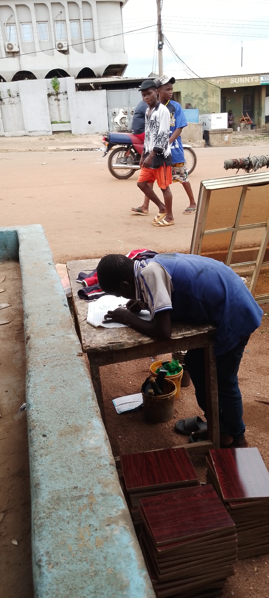 In Gboko, Nigeria, a young Black boy, wearing a blue vest over a white and blue striped shirt and dark pants, leans over a weathered wooden table, drawing on a piece of paper. His dark hair is visible as he focuses on his task. Scattered on the table are items of clothing, including a red, white, and blue striped garment and a pair of dark shorts. Beside the table, two yellow buckets and a dark bucket contain various objects, including green material and bottles. On the ground near the boy's feet are a pair of black sandals.
In the background, two young Black men are walking, one in a white shirt with a palm tree pattern and orange shorts, and the other in a blue shirt and patterned shorts. A motorcycle, partially visible, is parked behind them. Further in the distance, a pale building with arched windows and air conditioning units stands prominently. Across the street, a green building with the word "SUNNYS" painted on it is visible, with a person sitting outside. The scene is set on a dusty, unpaved road with scattered debris. Stacks of dark red, wood-patterned tiles are arranged to the right of the boy.