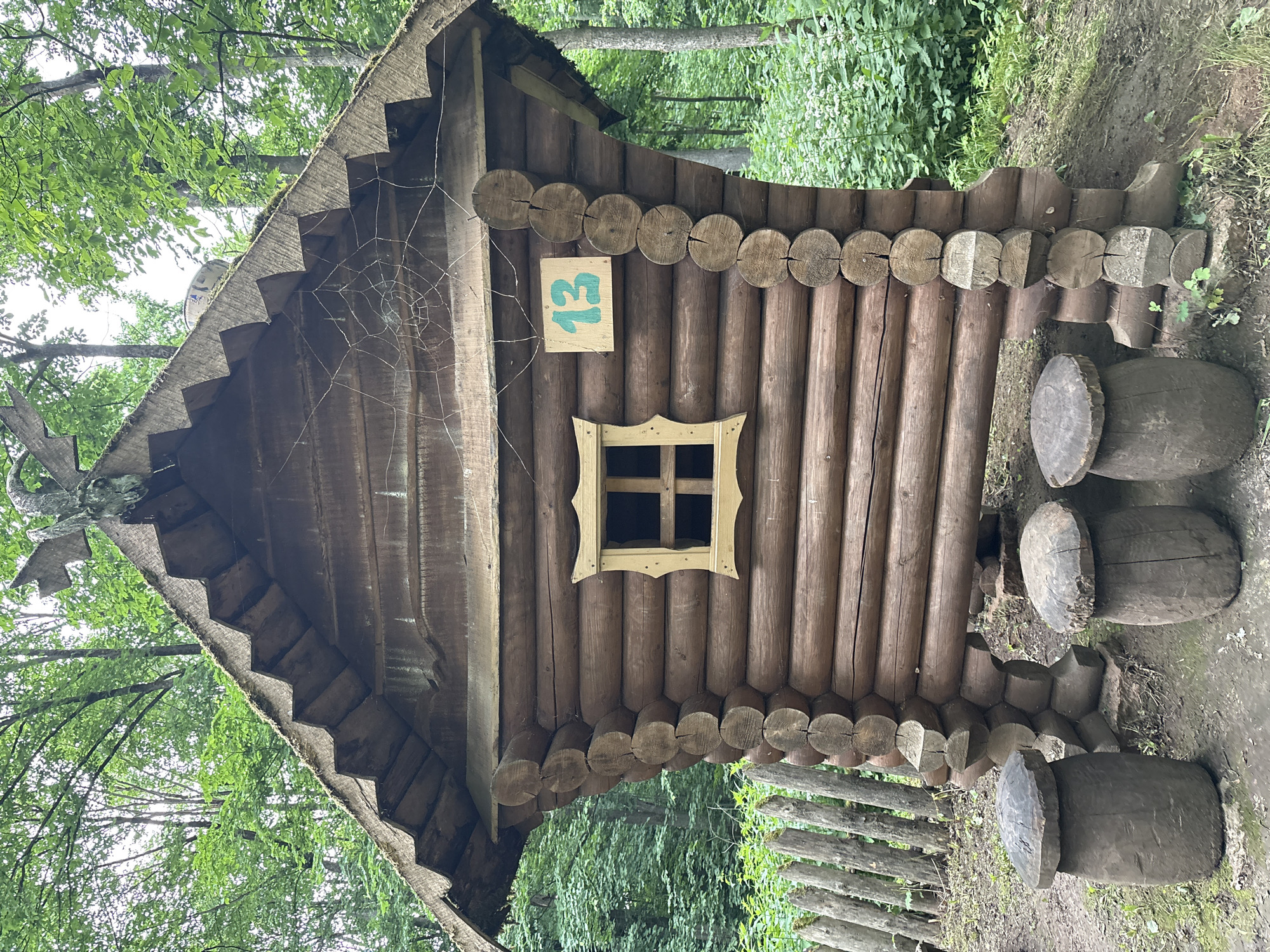 A rustic wooden structure resembling a small cabin or hut stands in a wooded area. The cabin is constructed from large, round logs and features a thatched roof. A small, square window with a decorative wooden frame is visible on the front of the cabin. A sign with the number "13" painted in green is affixed to the side of the cabin. In front of the cabin, three mushroom-shaped wooden stools are placed on the ground. The background consists of lush green trees and foliage, indicating an outdoor, natural setting. A rustic wooden fence is visible to the left of the cabin. The overall impression is that of a whimsical or fairytale-themed installation, possibly within a park or recreational area. The location is identified as Gor'ki, Russia.