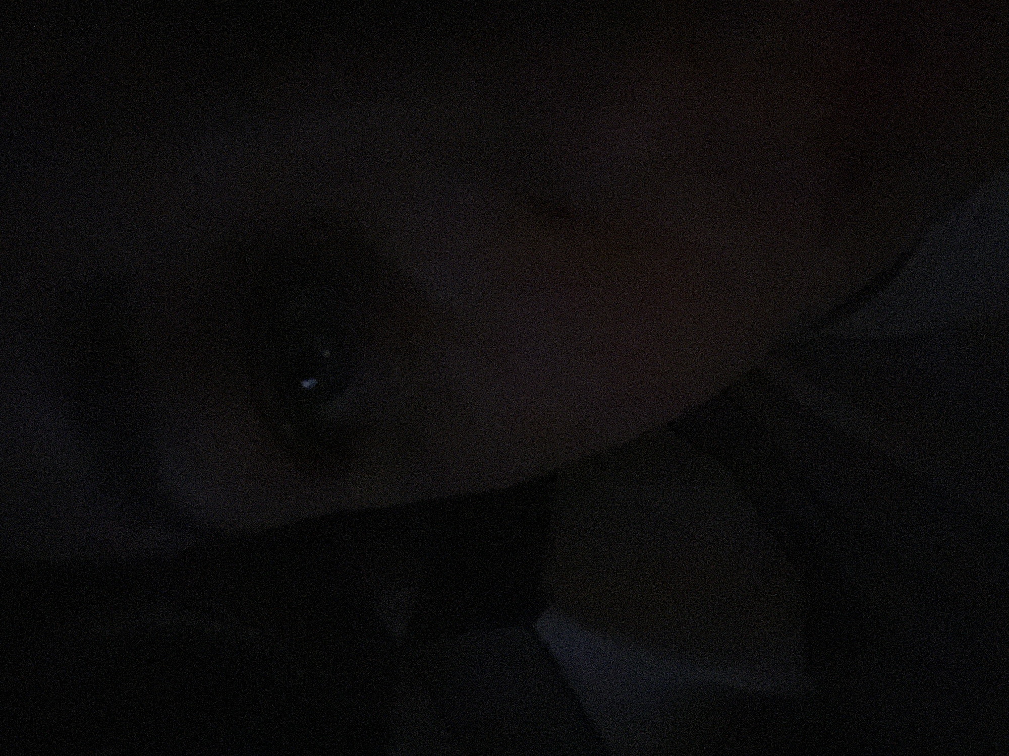 The image appears to be a close-up of a person's face in very low light. Only the eye and some facial features are vaguely discernible. The surrounding environment is dark and indistinct, with some blurry shapes suggesting fabric or clothing. There is insufficient information to determine a location or specific details about actions or interactions. The city of Chaikovsky, Russia, cannot be confirmed based on this image.