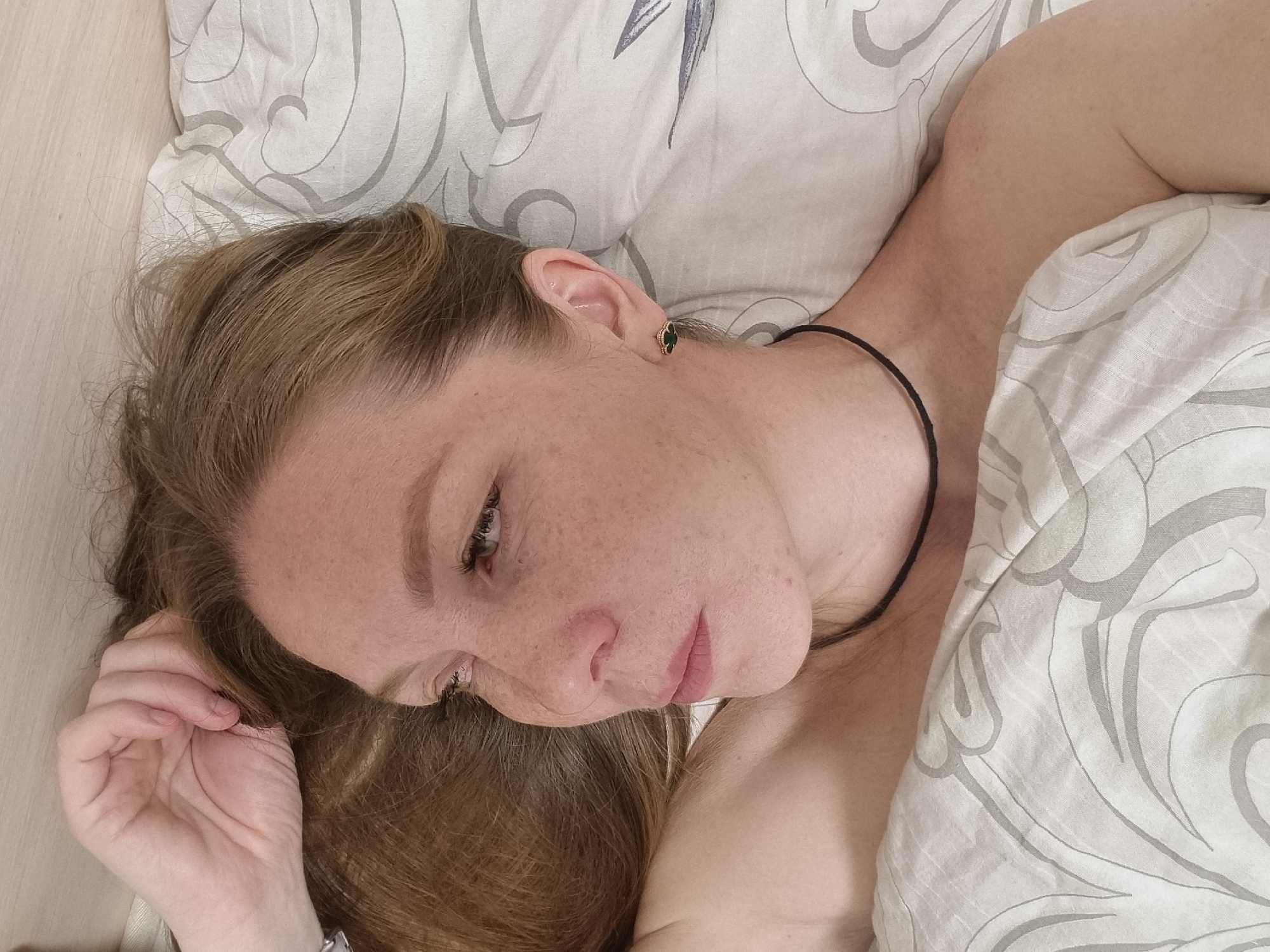 A light-skinned female with freckles and auburn hair is lying down, possibly in bed. She is on her back, with her head tilted slightly to the left. Her right hand is near her head, and she is wearing a thin black necklace. A green and gold earring is visible in her left ear. She is partially covered with a light-colored patterned blanket. The background includes a light-colored surface, possibly a wall or a headboard. No information suggests the scene takes place in Chaikovsky, Russia.